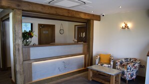 A reception area with a wooden reception desk, a small bench, a floral-patterned chair, and a wall-mounted clock. - White Hart Inn (Alfreton)