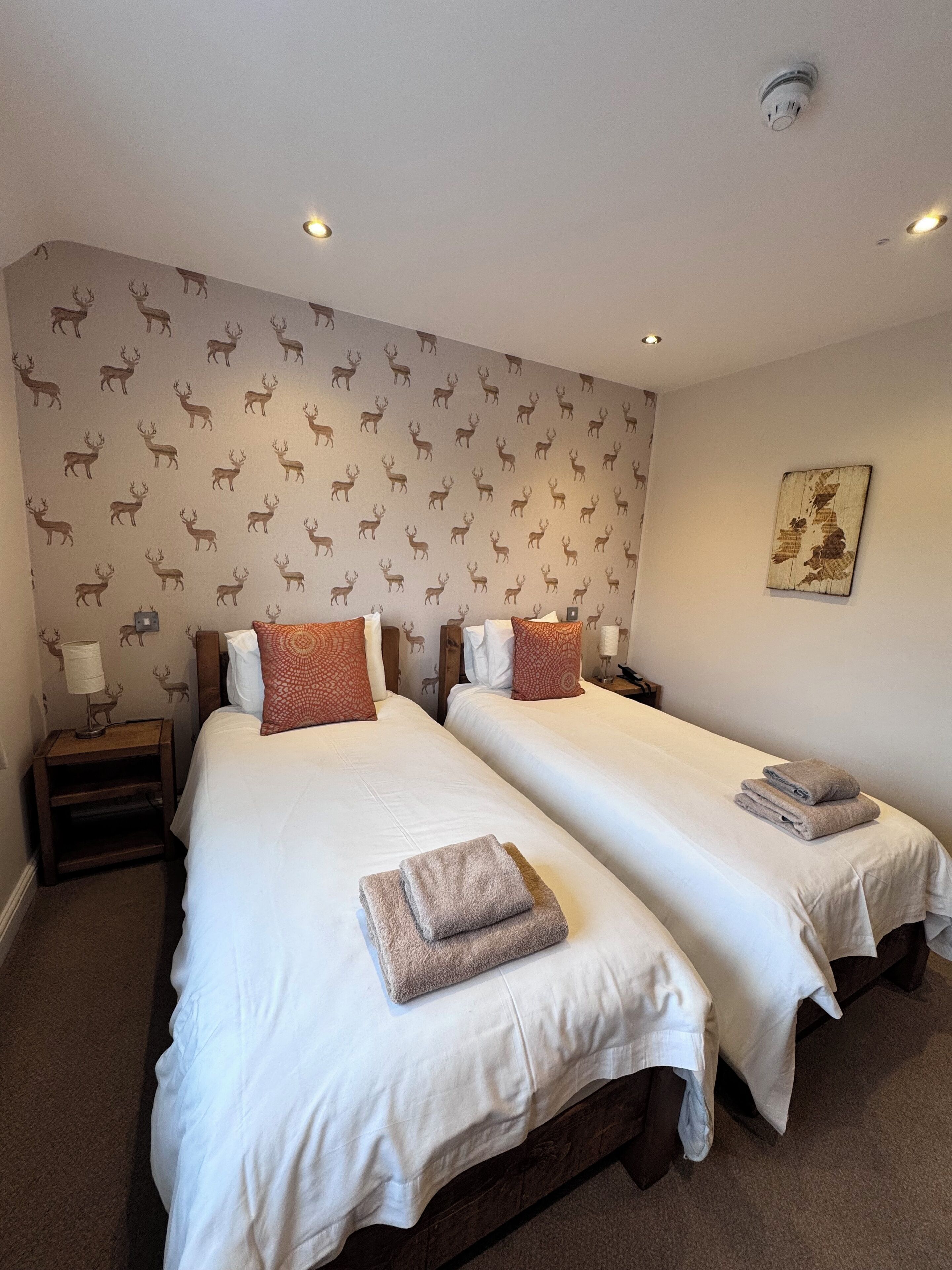A hotel room with two single beds, each with a pillow and a folded towel. The room has a patterned wallpaper and a small table with a lamp.