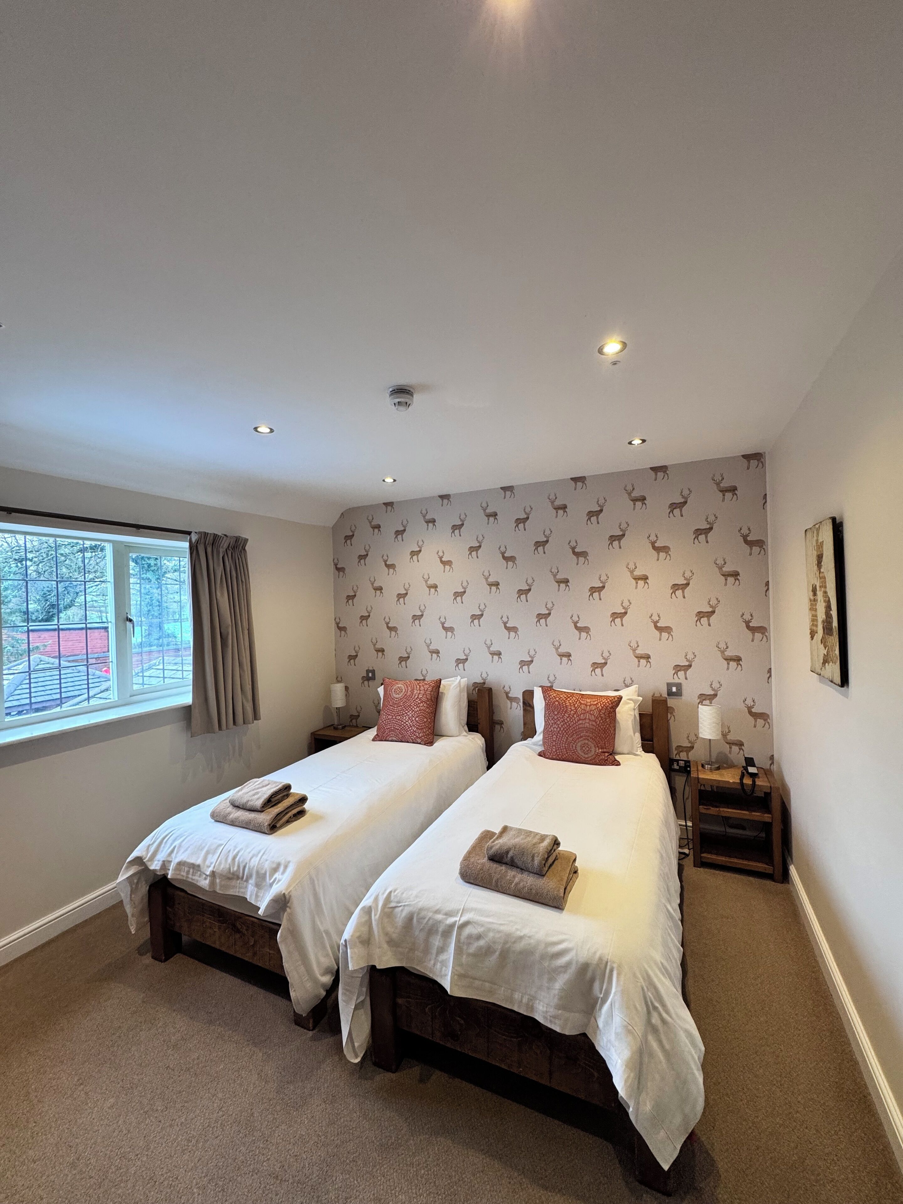 A hotel room with two beds, a window with curtains, a patterned wallpaper, and a small table with a lamp.
