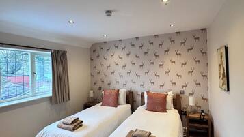 A hotel room with two beds, a window with curtains, a patterned wallpaper, and a small table with a lamp.