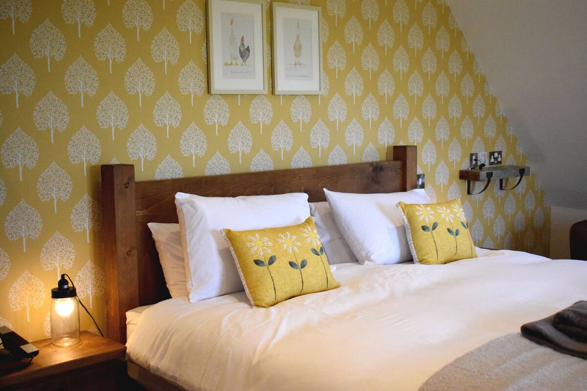 Cama bem arrumada com almofadas brancas e amarelas, cabeceira de madeira e mesa de cabeceira com abajur.