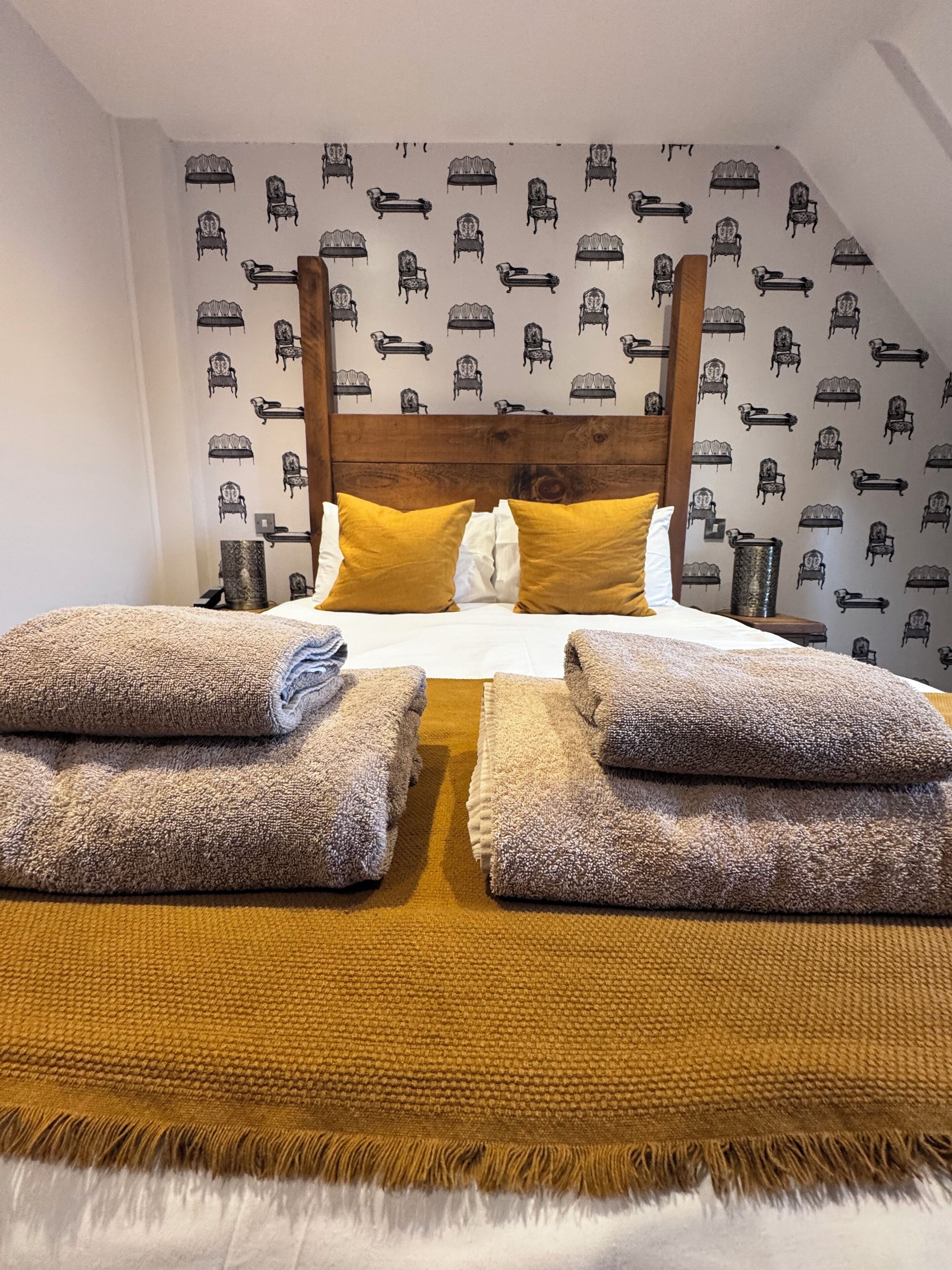 A bedroom with a wooden headboard, a bed with two pillows, and a patterned wallpaper.