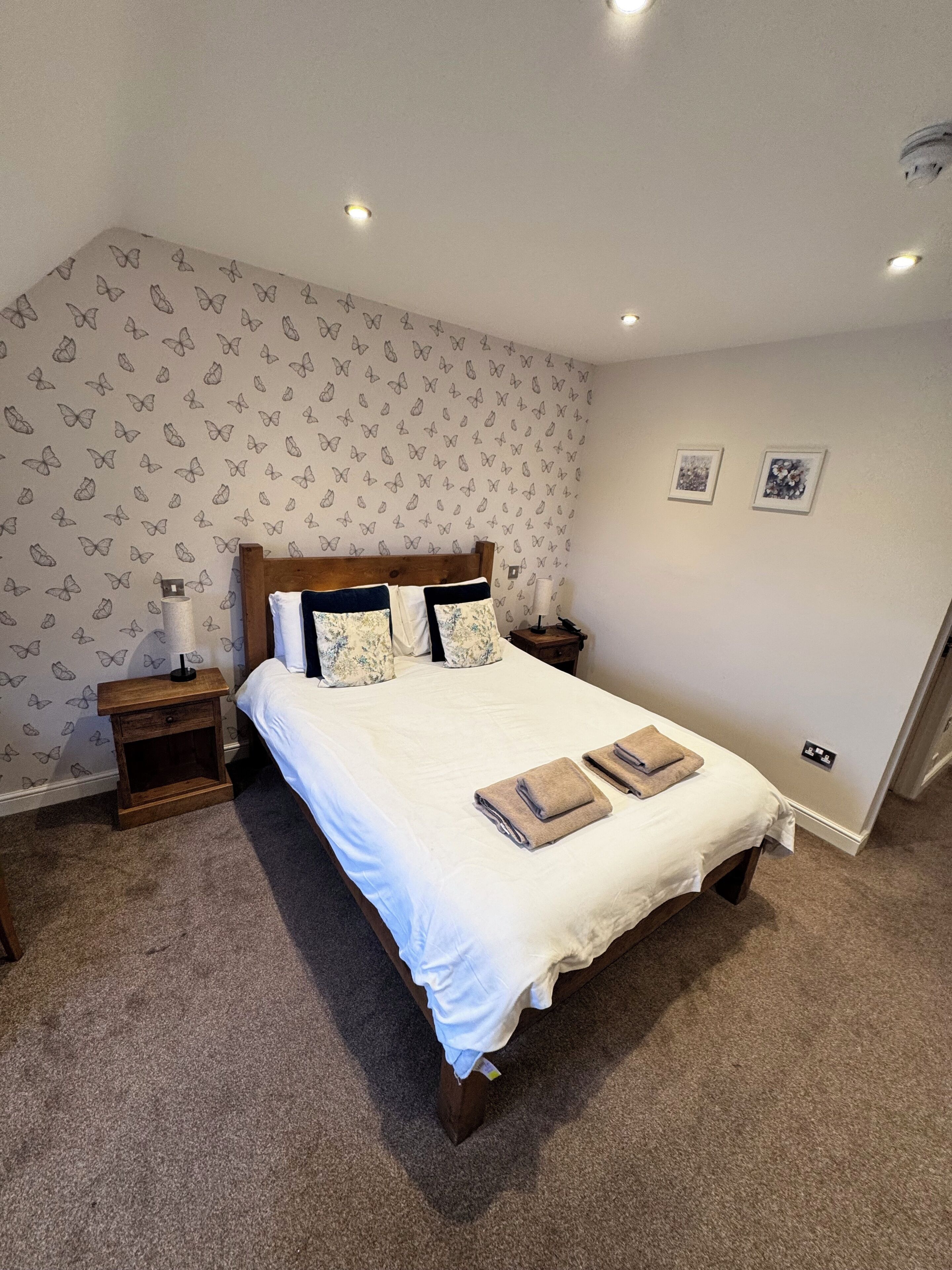 Una cama bien hecha con ropa blanca y dos almohadas, una mesita de noche de madera con una lámpara, y una pared con diseños de mariposas.