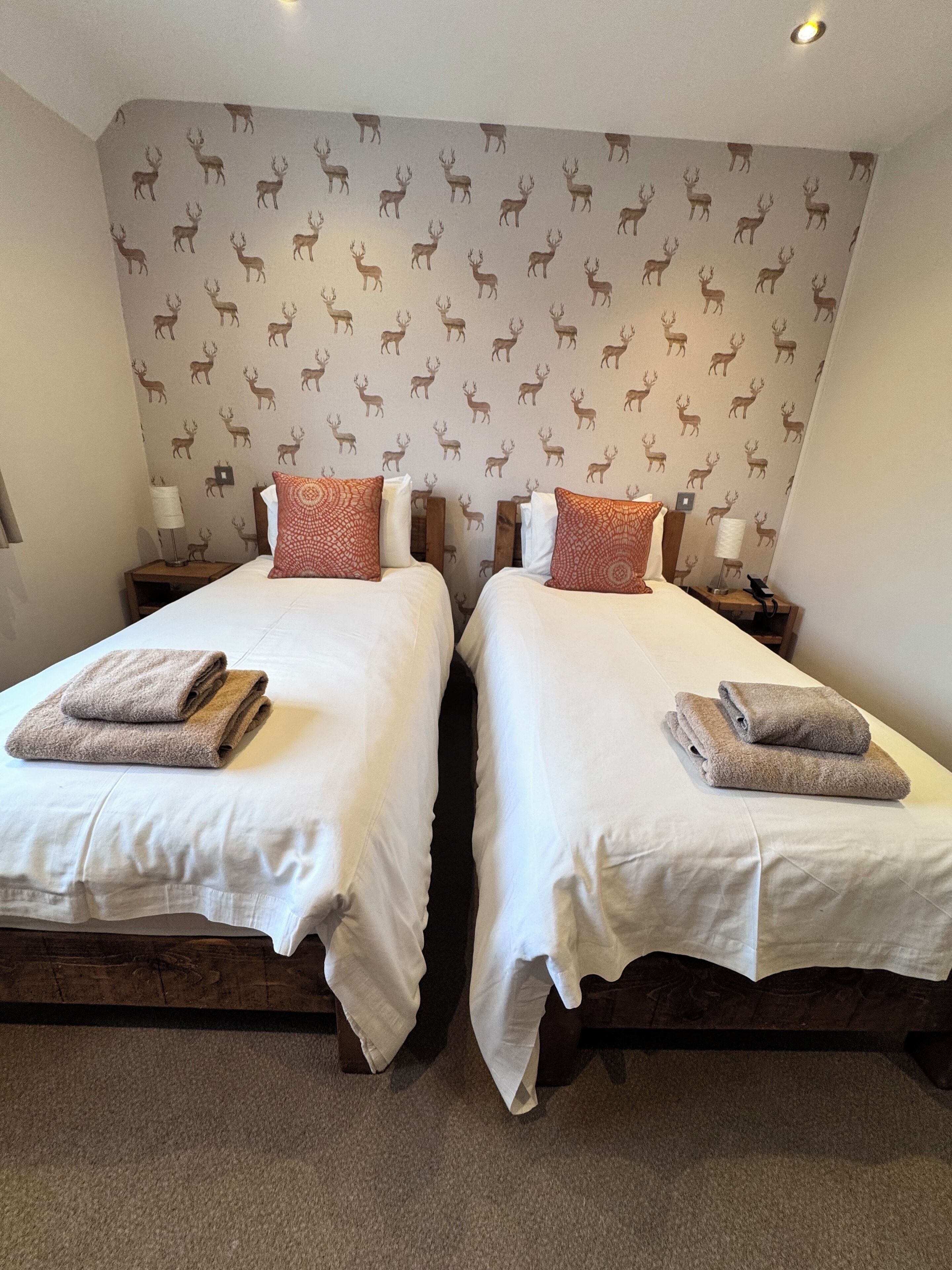 A room with two beds, each with white linens and brown towels. The walls have a deer pattern wallpaper.