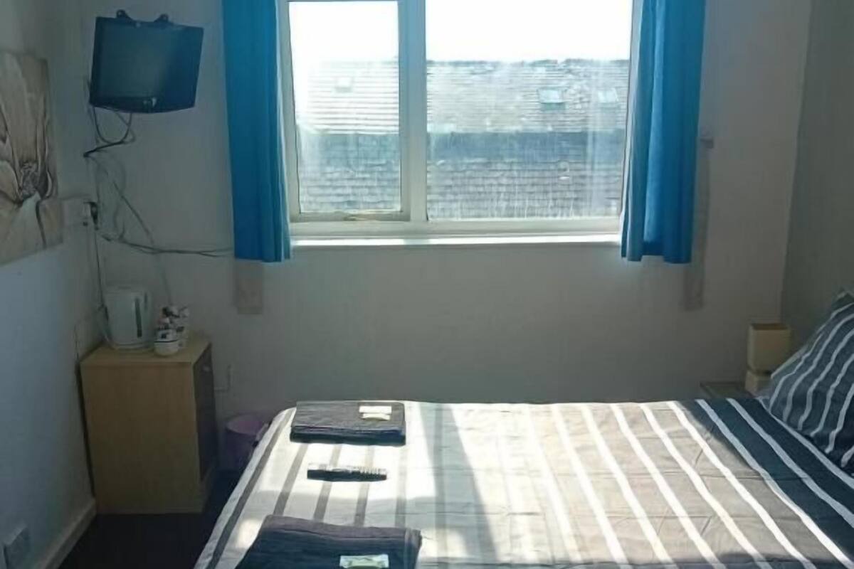 Un dormitorio con una cama, una ventana con cortinas, una lámpara de noche y un televisor en la pared.