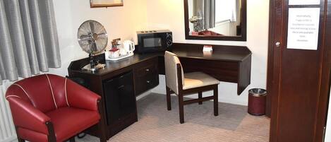 Habitación de hotel con escritorio, una silla roja, microondas, cafetera, un bote de basura y un cartel sobre la estación de planchado.