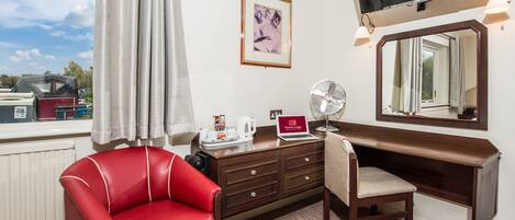 Ein Hotelzimmer mit einem roten Ledersessel, einem Holztisch, einem Laptop, einem Ventilator, einem Fernseher und einem gerahmten Bild an der Wand.