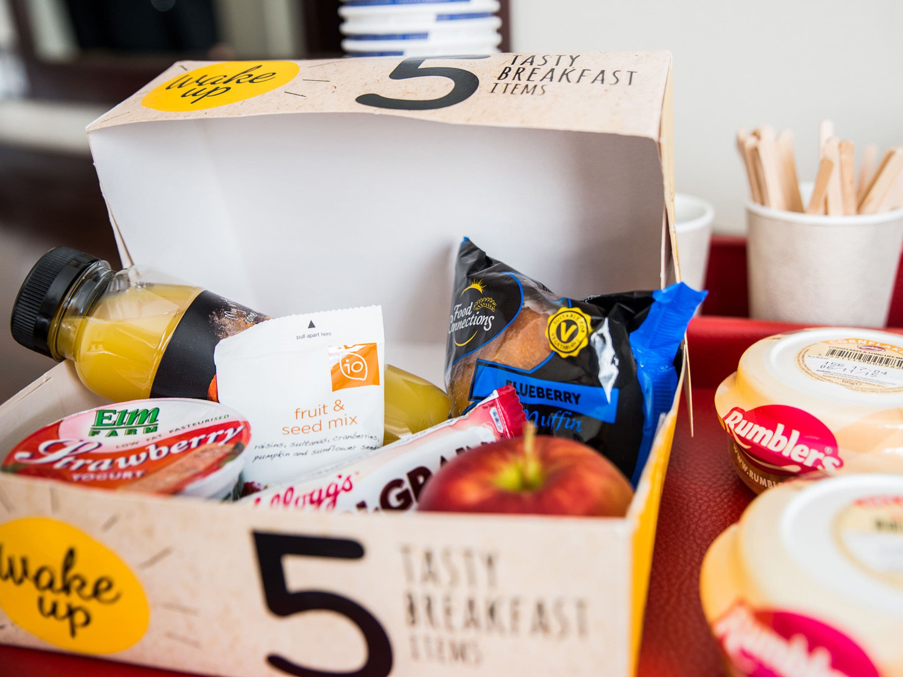 A breakfast box labeled '5 TASTY BREAKFAST ITEMS' containing a variety of food items including yogurt, cereal, a fruit & seed mix, and a muffin.