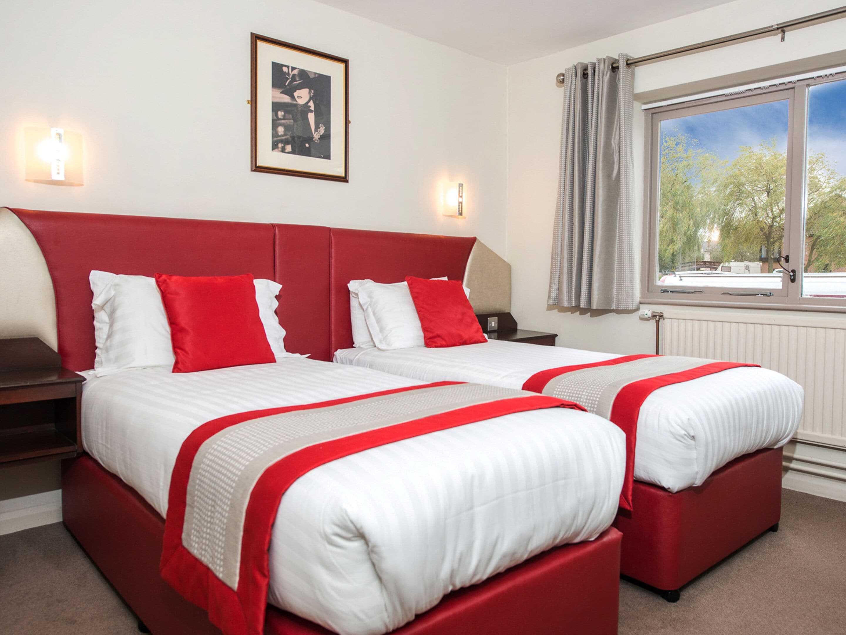 A hotel room with two beds, each with red and white bedding, a framed picture on the wall, and a window with curtains.