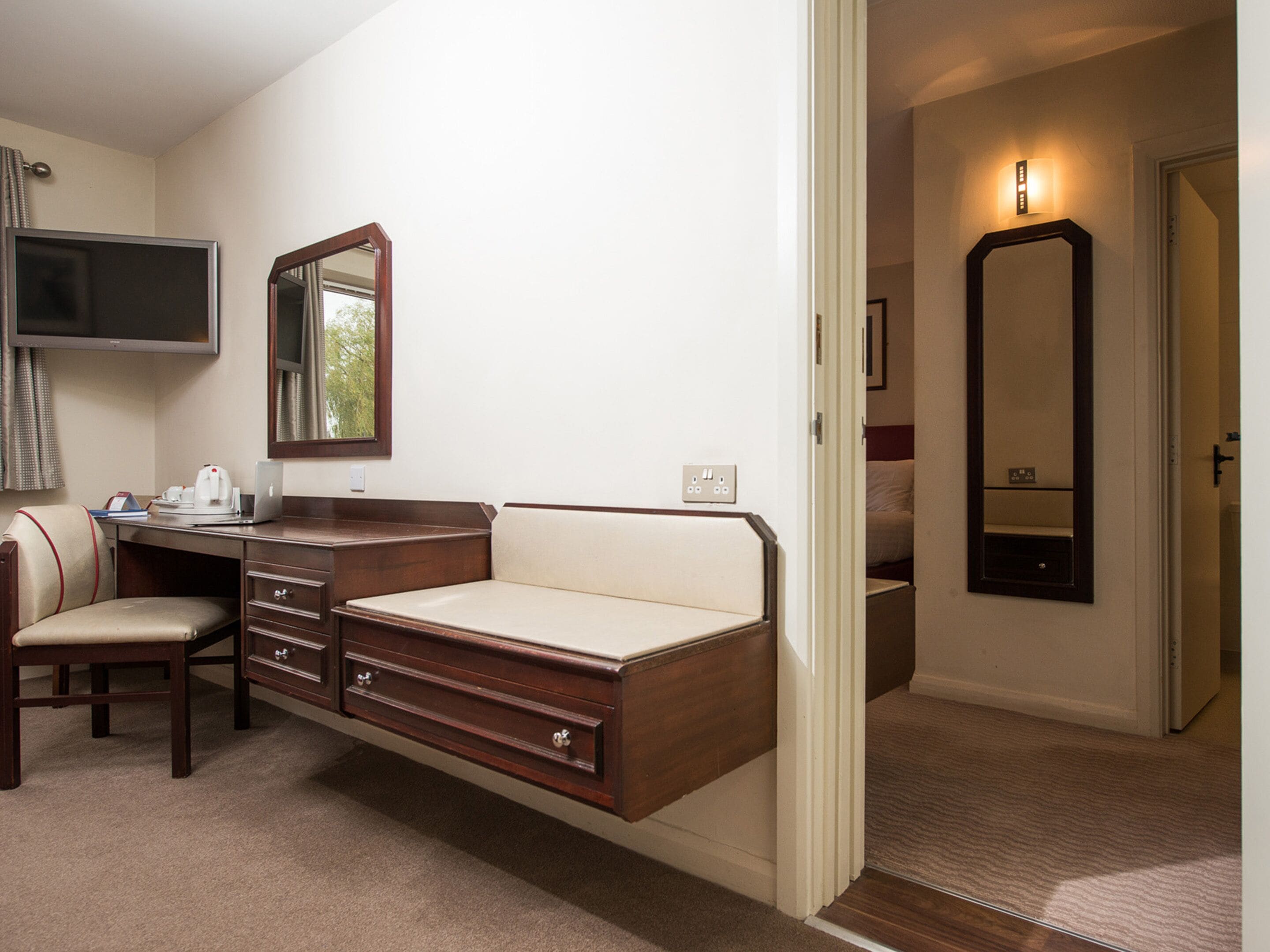 A hotel room with a desk, chair, and a bed. There is a television on the wall and a mirror above the bed.