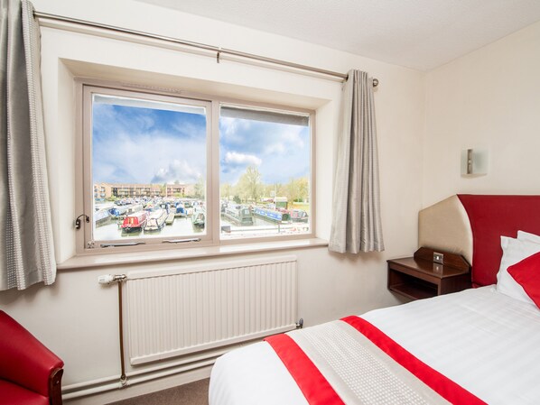 Habitación de hotel con un ventanal grande, una cama, una silla roja, una mesita de noche y vistas a un canal con barcos.