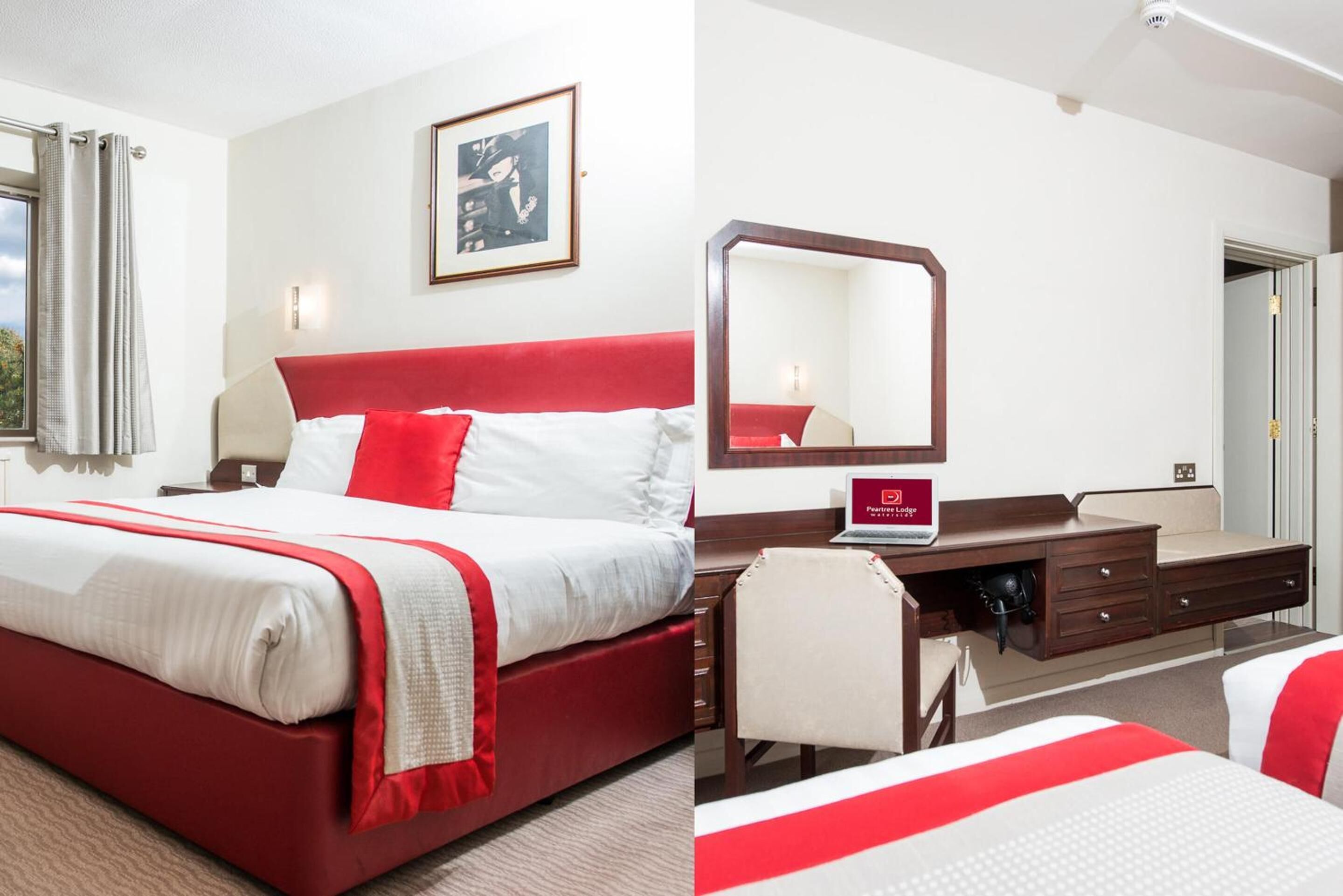 Habitación de hotel con una cama roja y blanca, un escritorio con una computadora portátil, un espejo y un cuadro en la pared.