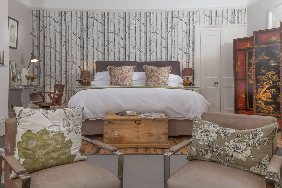 Quarto com cama, duas cadeiras, mesa de cabeceira, abajur e parede com padrão de árvore.