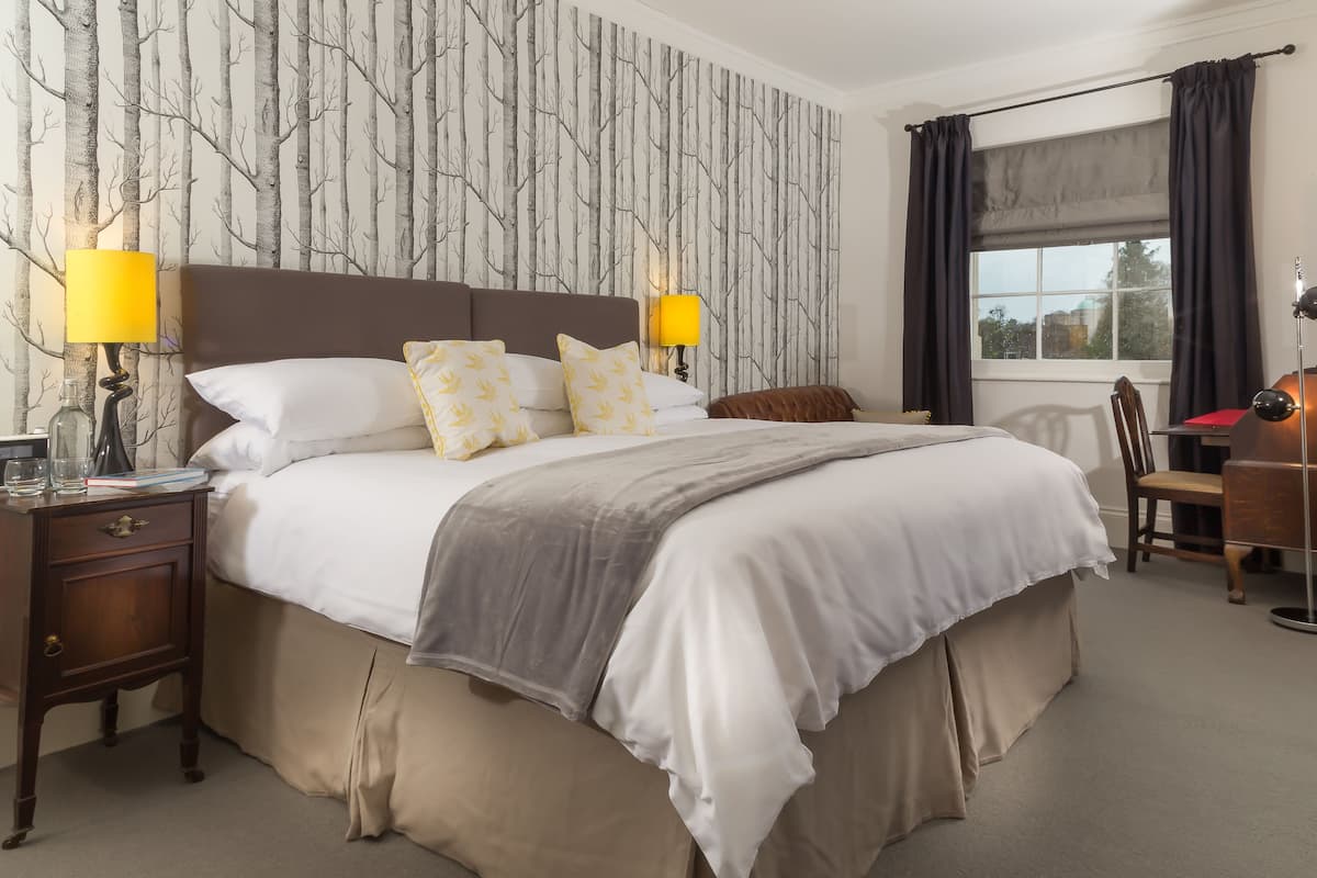Uma cama bem arrumada com cabeceira marrom, duas luminárias de apoio e uma janela com cortinas escuras.