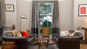 A living room with two leather sofas, a wooden coffee table, a floor lamp, and a view of greenery outside. - Ravenhurst Bed and Breakfast (Cheltenham)