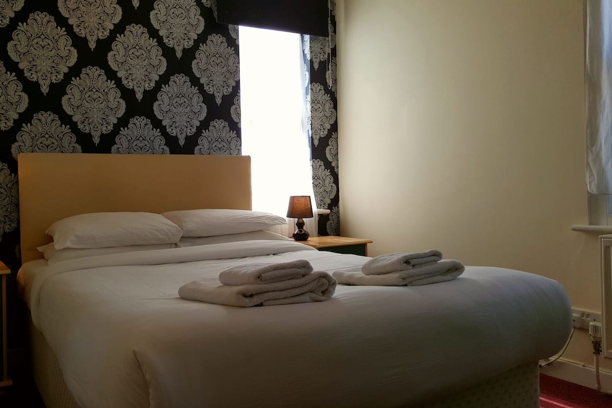 A neatly made bed with white linens and towels, two bedside lamps, a nightstand, and a patterned wallpaper.