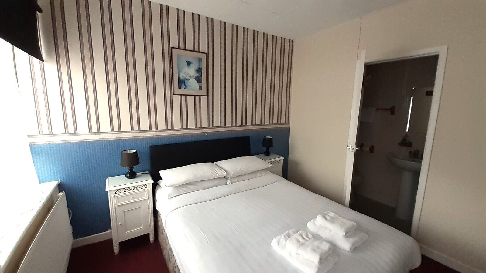 Habitación de hotel con cama, dos mesitas de noche, una mesita auxiliar, un cuadro enmarcado en la pared y un baño visible a través de una puerta abierta.