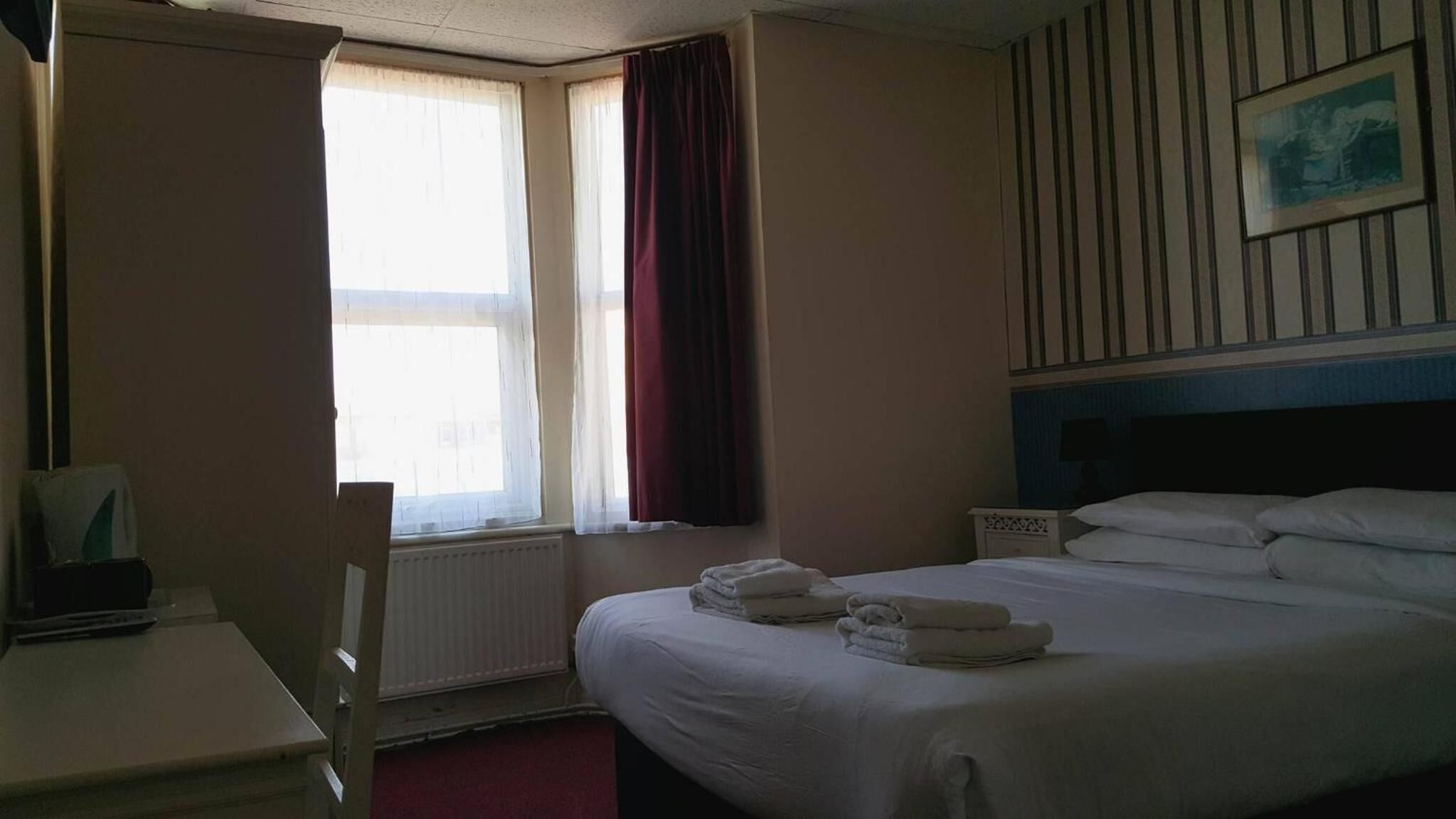 A hotel room with a bed, a desk, a chair, a window with curtains, and a framed picture on the wall.