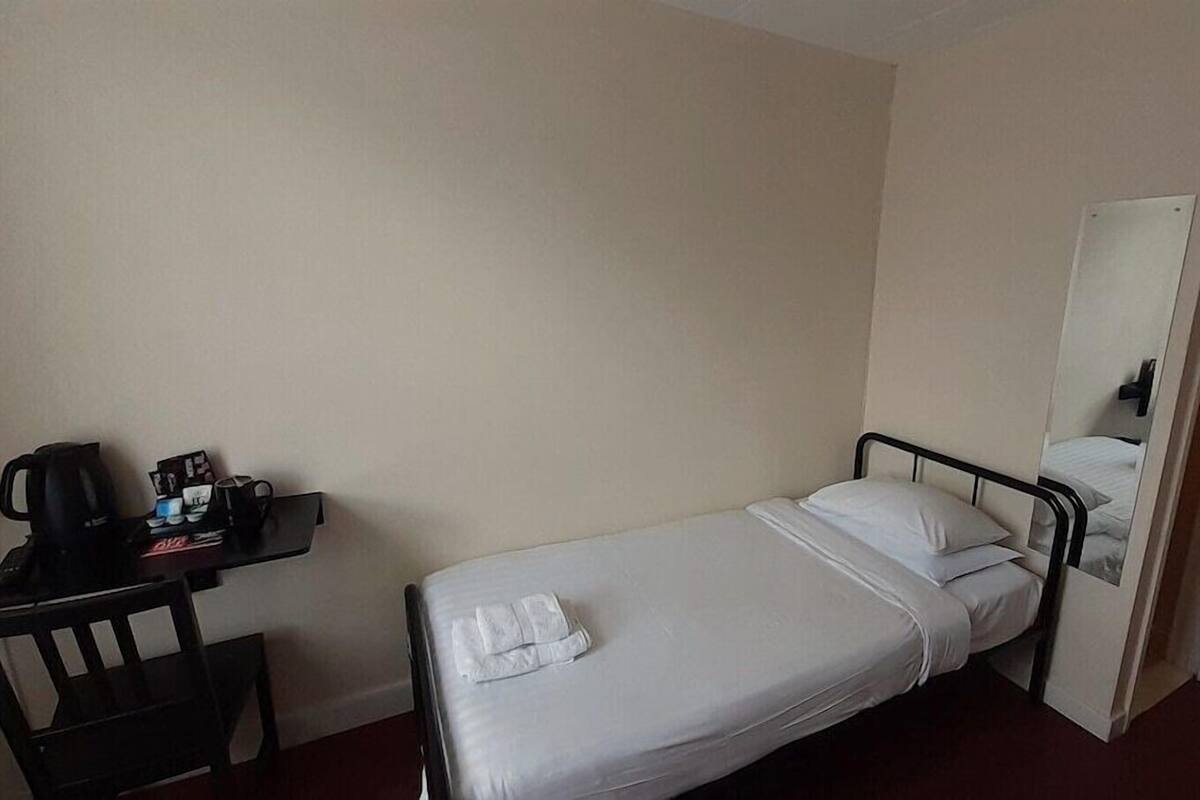 A single bed with white linens, a small desk with a black chair, a coffee maker, and a mirror on the wall.