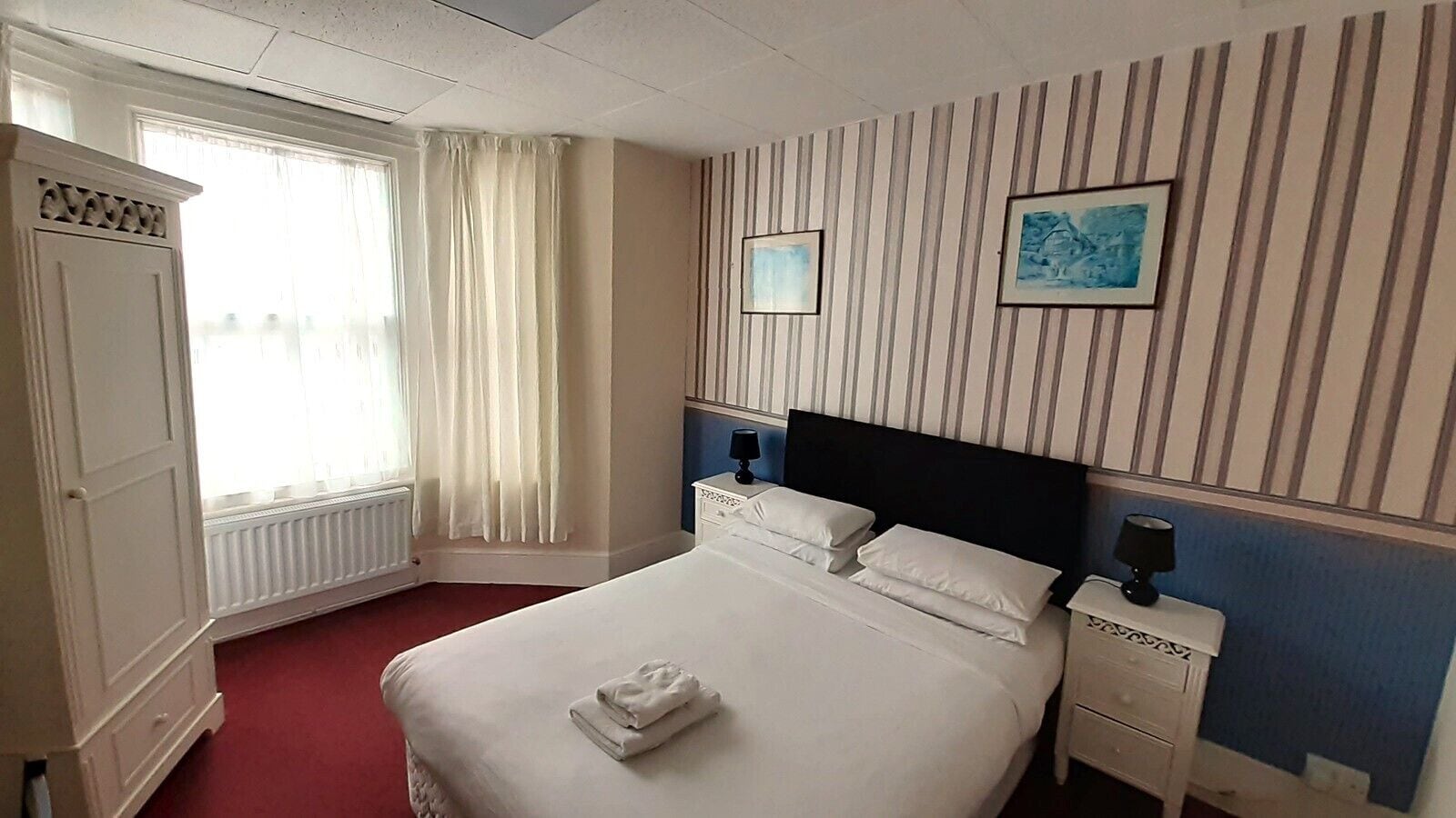 A hotel room with a large bed, a nightstand, a window with curtains, and a striped wall.