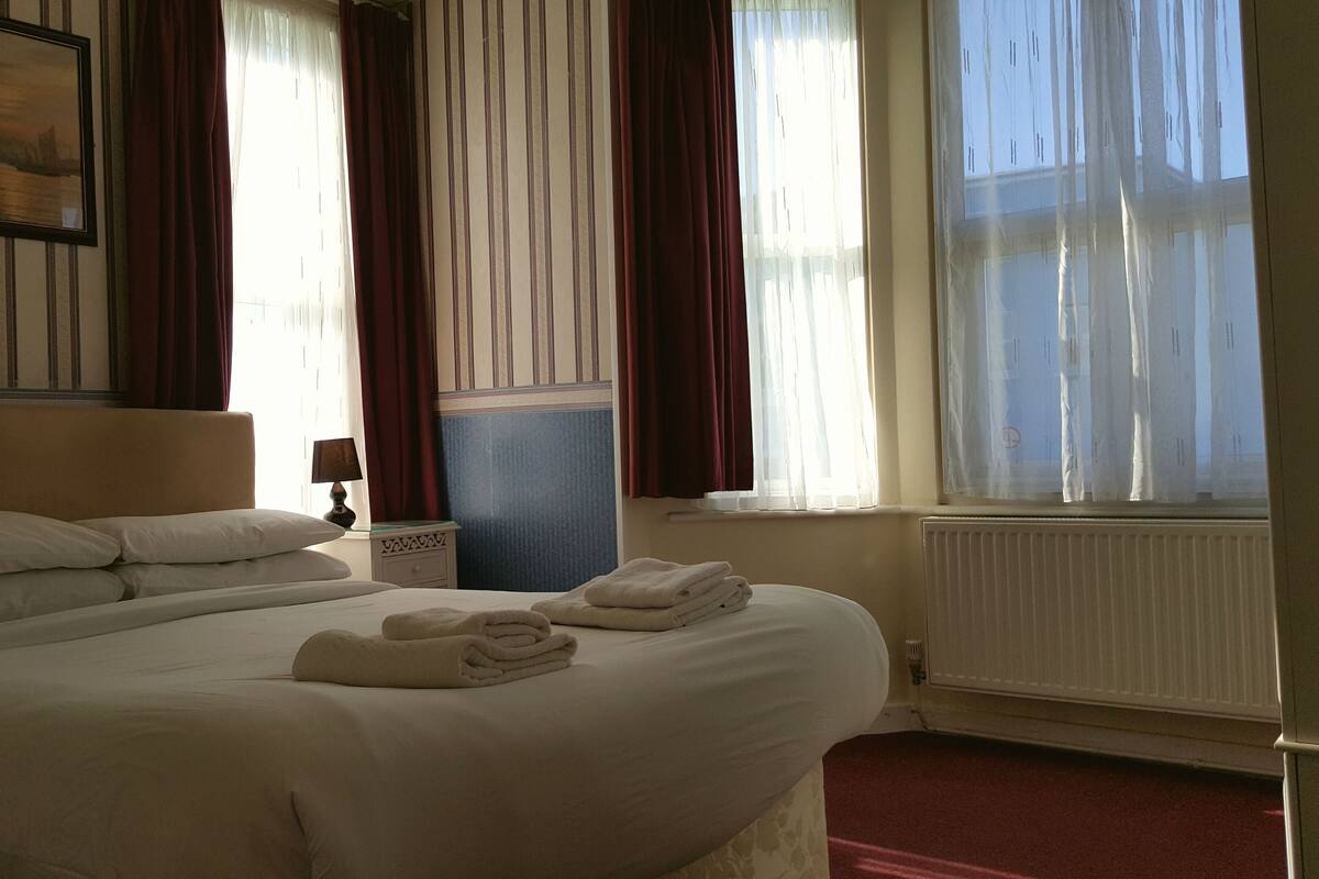 A hotel room with a large bed, a nightstand, a lamp, a radiator, and a window with curtains.
