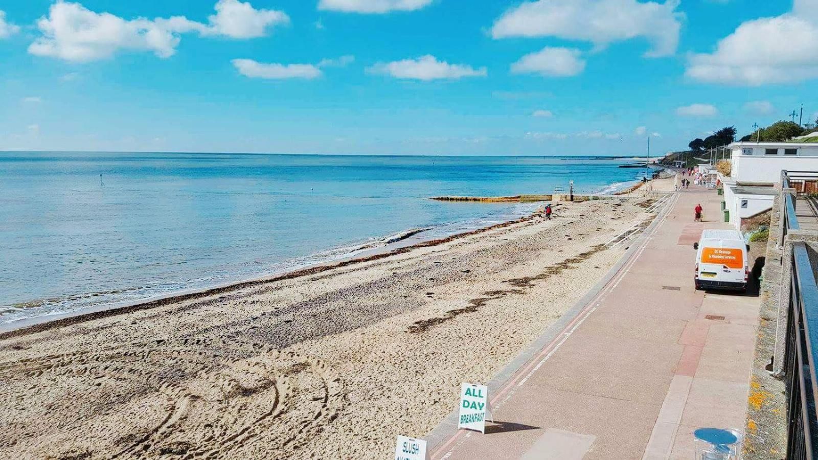 A beach with a paved walkway, a sign indicating 'ALL DAY BREAKFAST', and a van parked on the side.