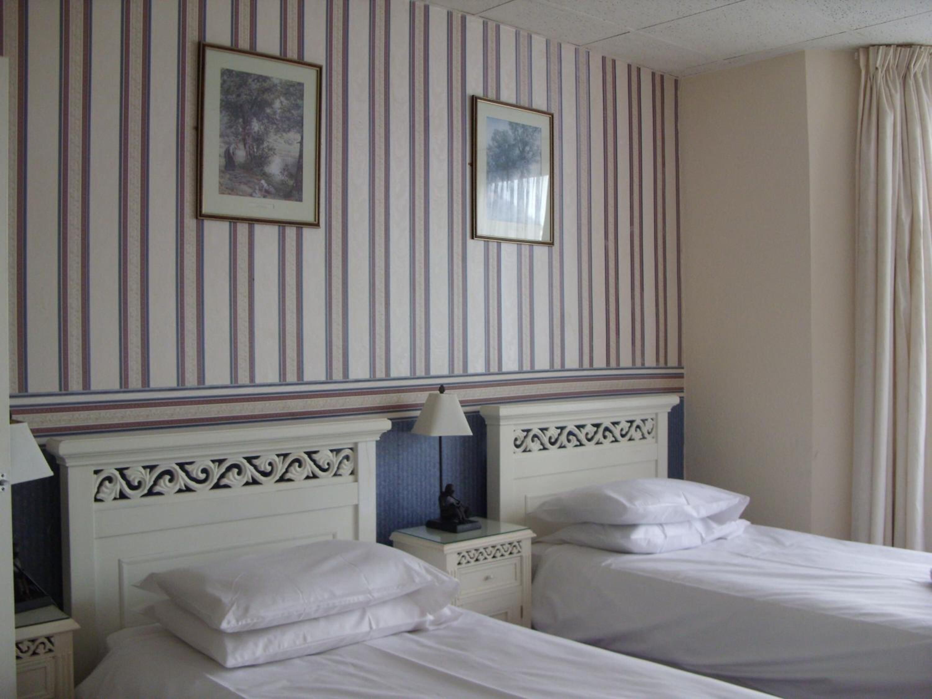 Une chambre d’hôtel avec deux lits, un papier peint à rayures, des tableaux encadrés et une table de chevet avec une lampe.