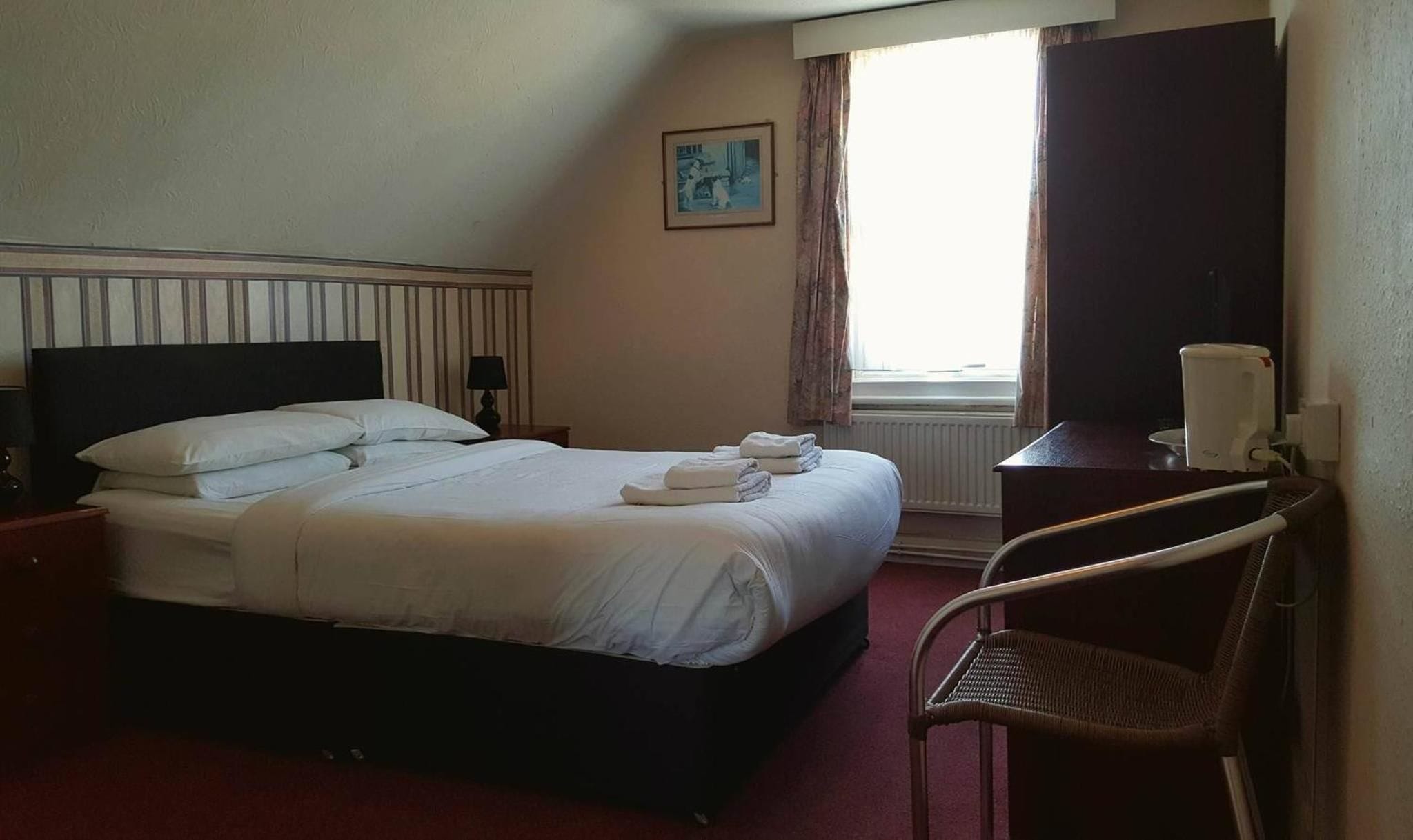 Ett hotellrum med en säng, en stol, ett skrivbord, ett fönster med gardiner och en tavla på väggen.