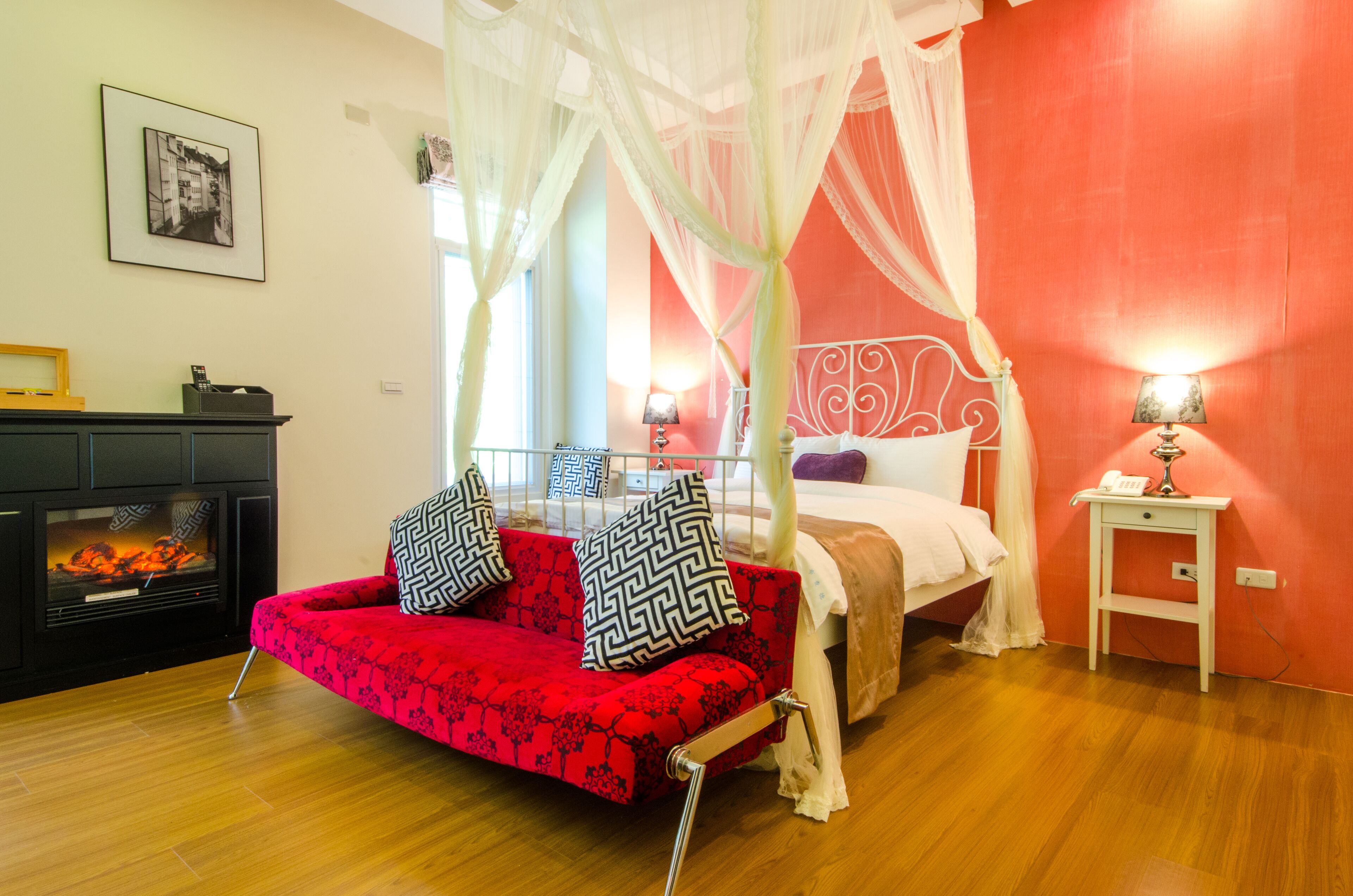 A bedroom with a red sofa, a canopy bed, a bedside table with a lamp, and a fireplace.