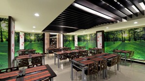 A restaurant with multiple tables and chairs, a ceiling with black slats, and large forest-themed murals on the walls.