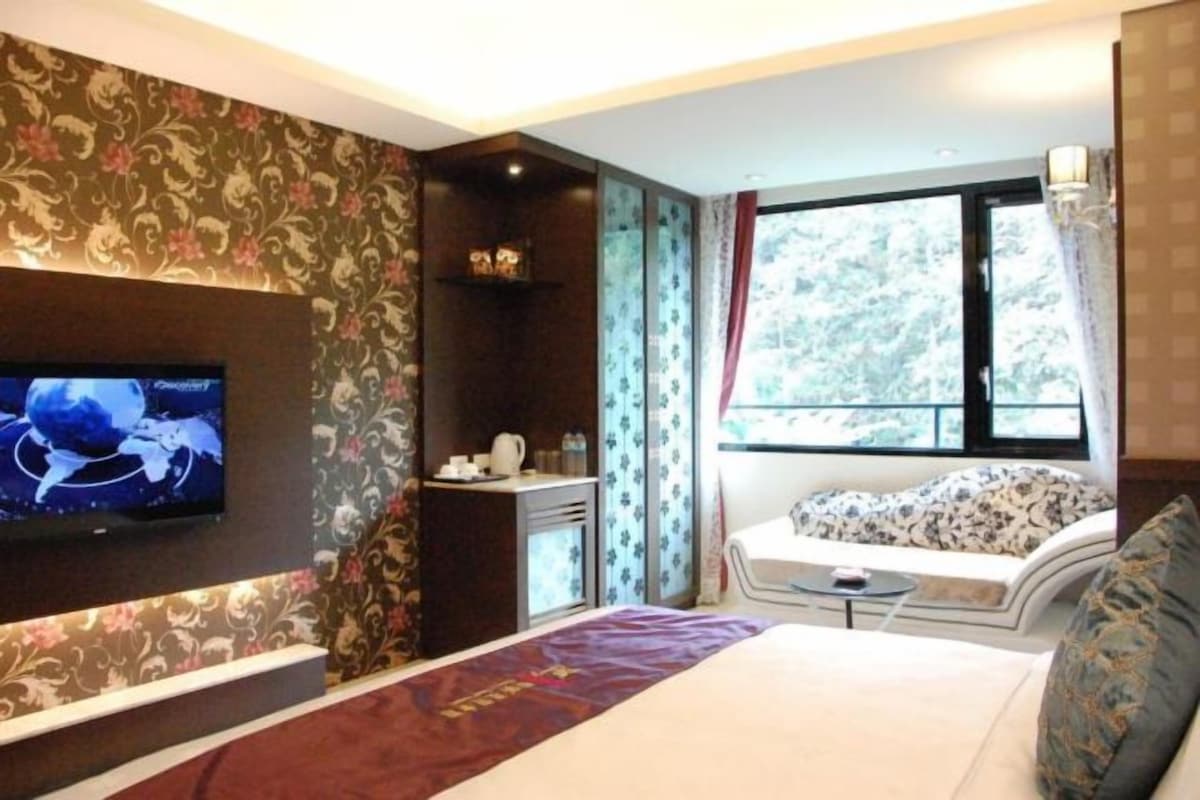 Quarto de hotel com cama, televisão na parede, uma mesinha e janela com vista para árvores.