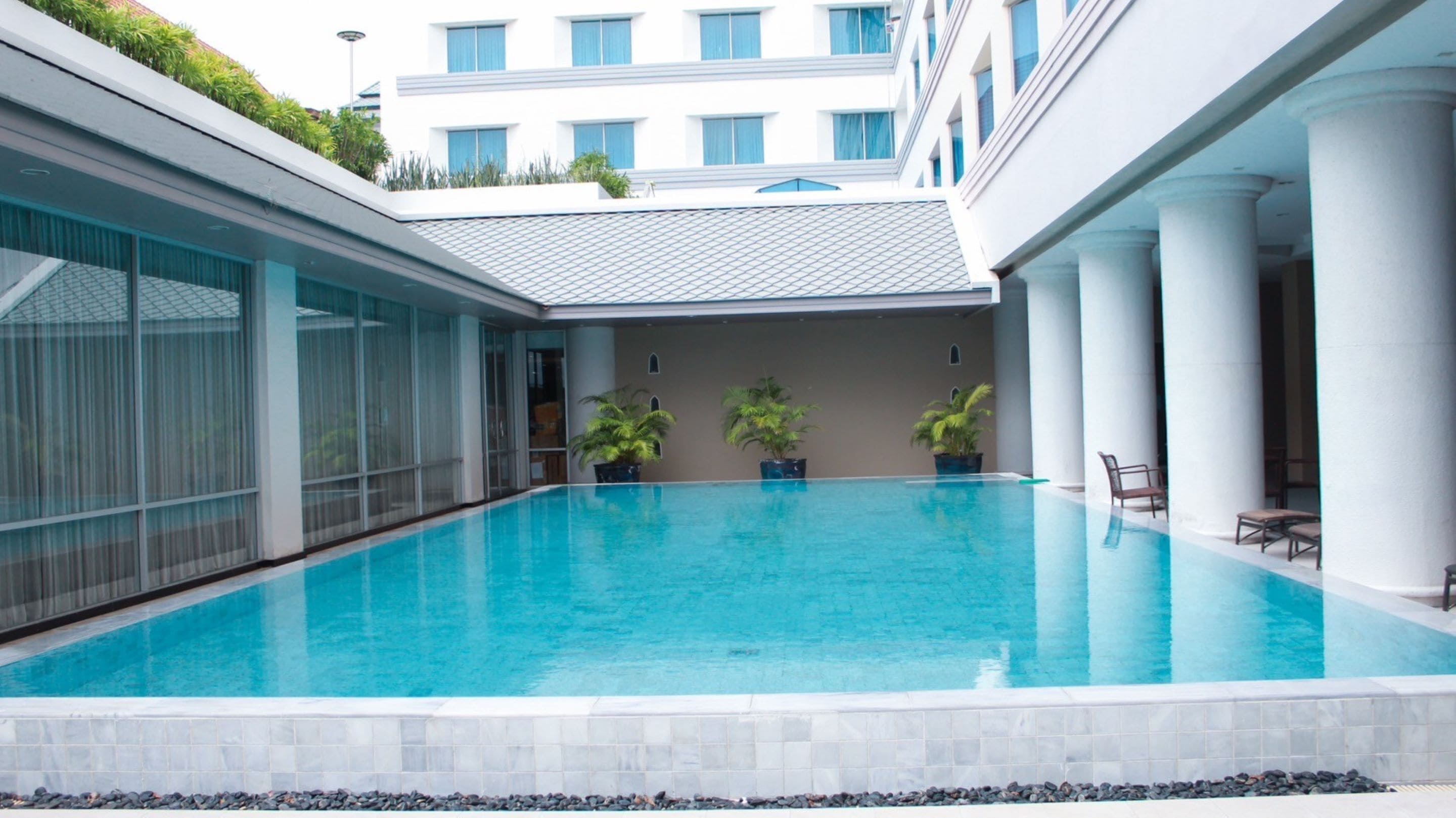 A modern indoor swimming pool with clear blue water, surrounded by white columns and potted plants.