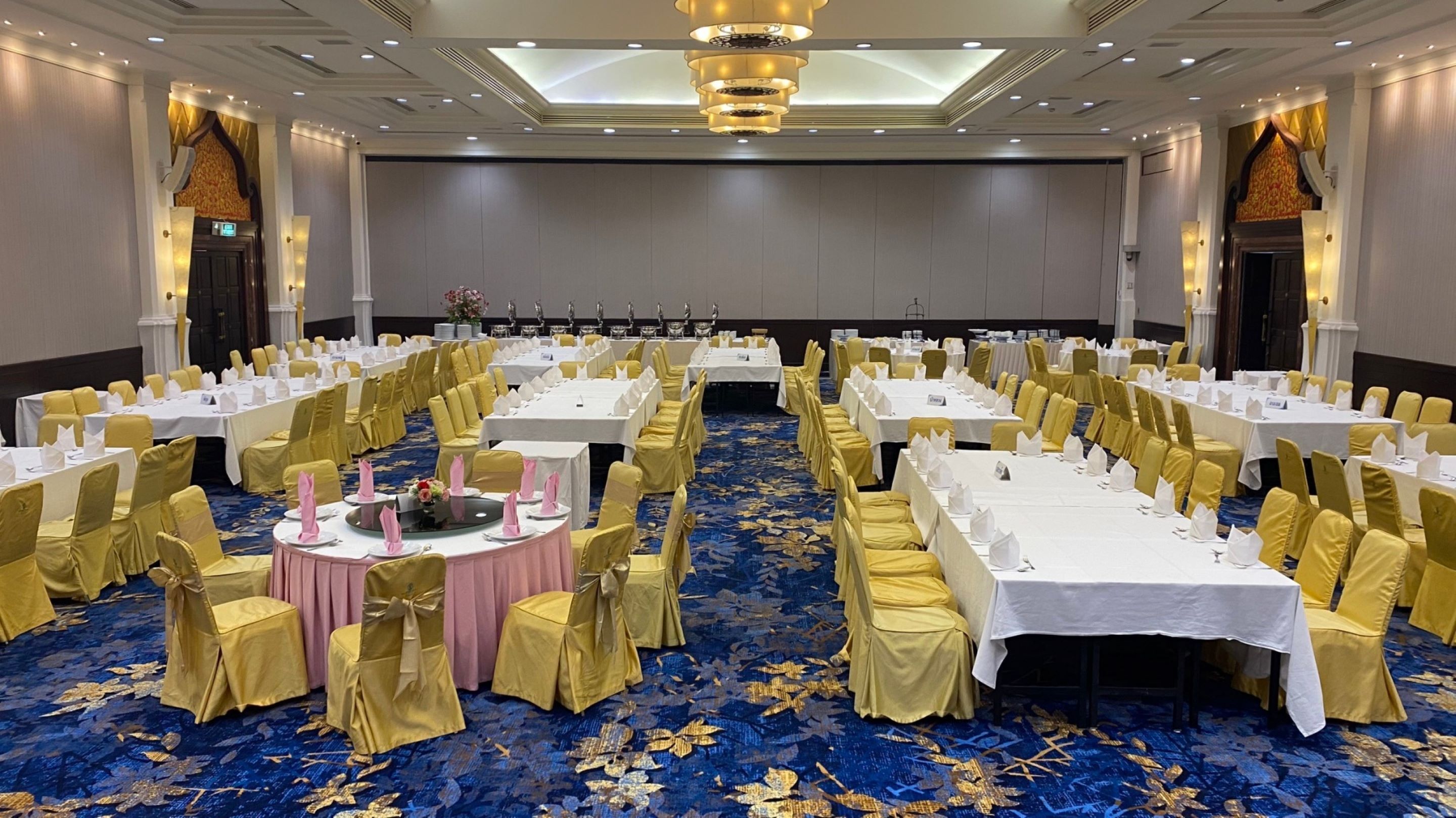 A banquet hall with round tables and chairs, a blue carpet with gold patterns, and a chandelier.