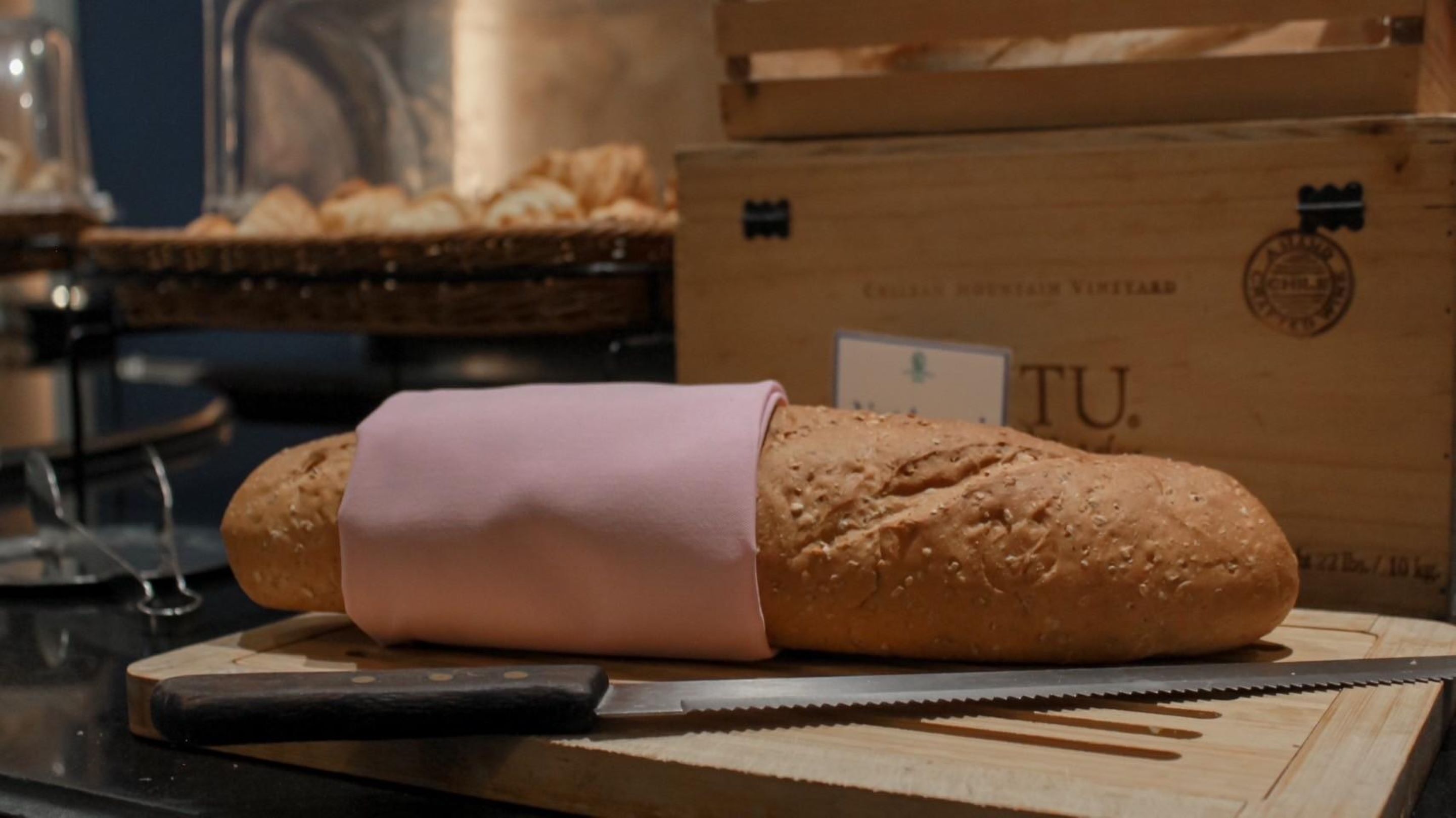 A loaf of bread with a pink wrapper on a wooden cutting board, a knife, and a cardboard box with 'TU' visible in the background.
