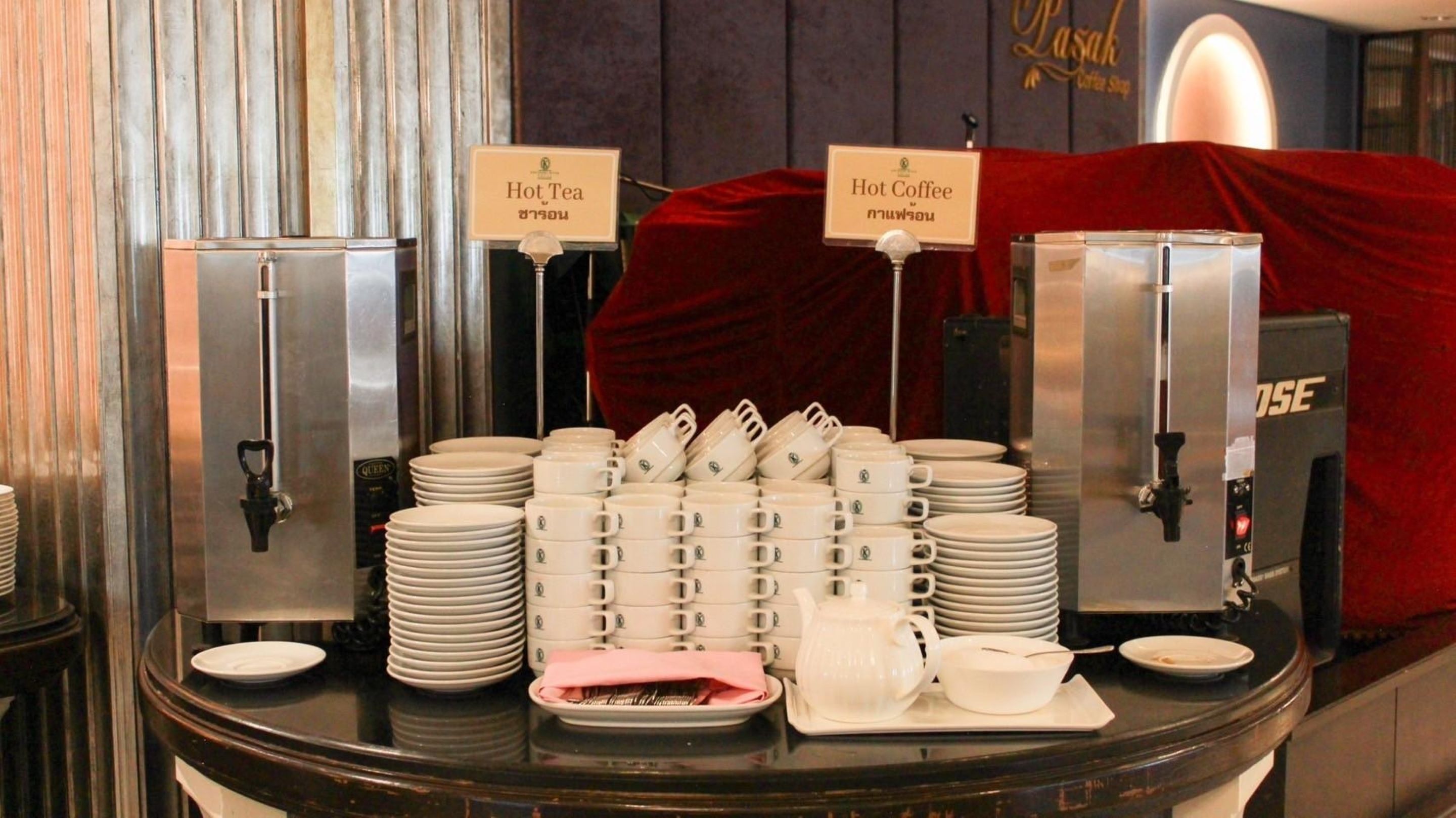 A buffet setup with hot tea and hot coffee dispensers, neatly stacked plates, bowls, and utensils.