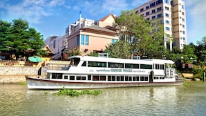 Un barco fluvial con un cartel que dice "Krungsri Princess" atracado cerca de un edificio con pared de piedra y árboles.