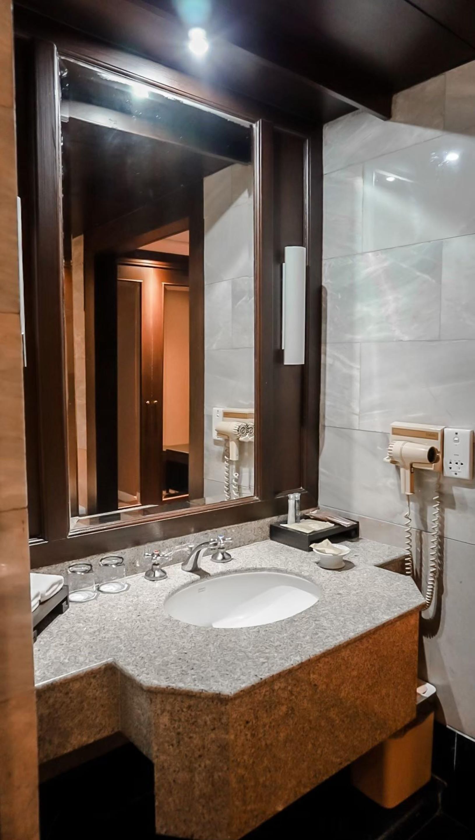 A modern bathroom with a granite countertop, a white sink, a large mirror, and a hairdryer mounted on the wall.