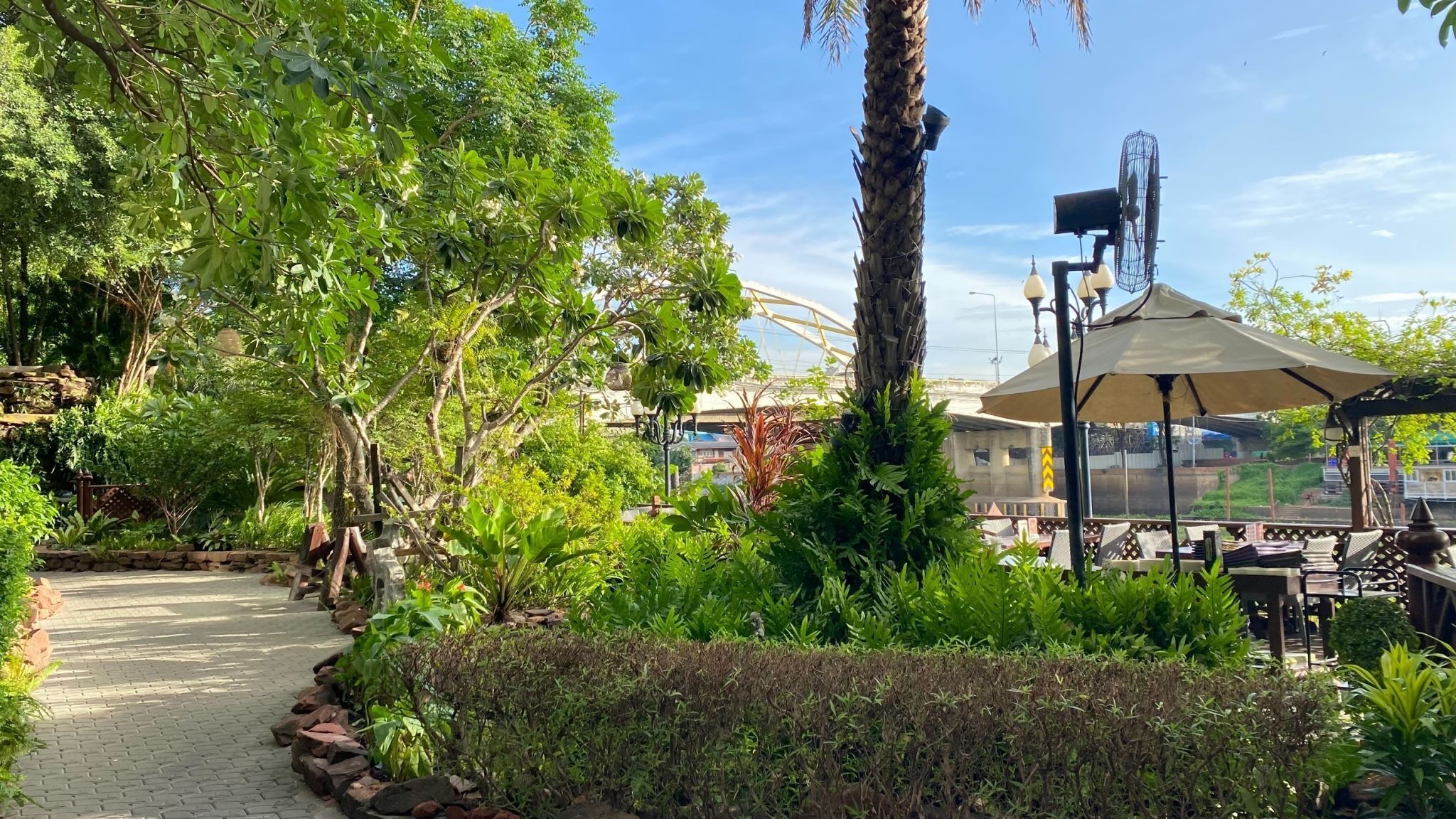 A garden pathway leading to a gazebo with outdoor seating, surrounded by lush greenery and a clear sky.