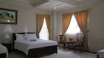 A hotel room with a bed, a chair, a small table with a vase of flowers, and a painting on the wall.