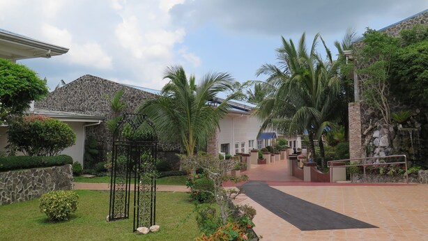 Un patio con muro de piedra, palmeras, una reja metálica y un camino que lleva a un edificio con techo azul.