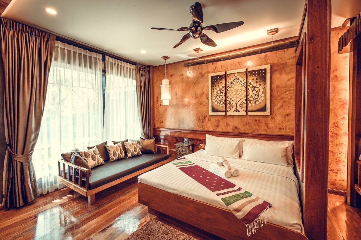 A spacious bedroom with a large bed, a sofa bed, wooden flooring, a ceiling fan, and a decorative wall panel.