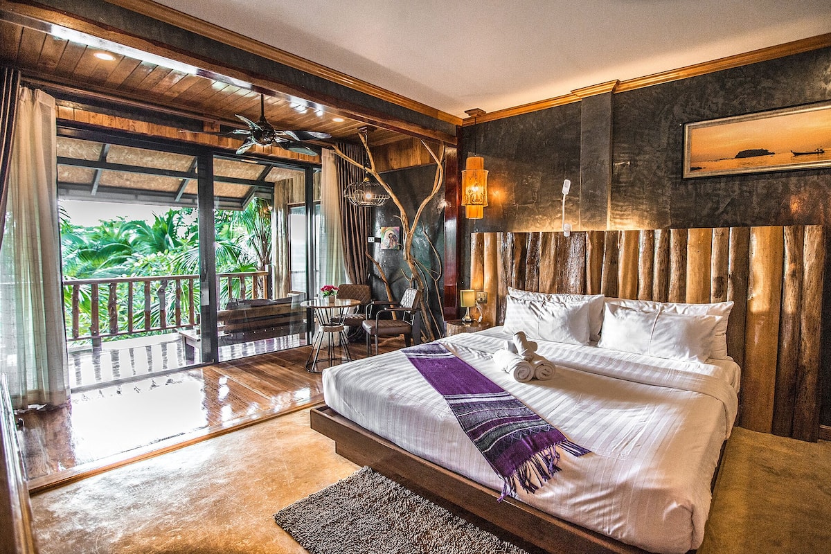 A spacious bedroom with a large bed, a balcony with a view of greenery, and a wooden ceiling.