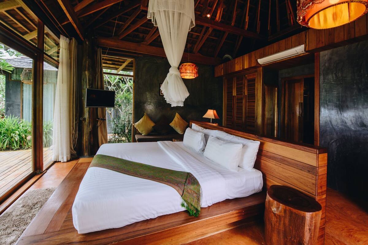 A wooden-framed bedroom with a bed, a TV, and a view of greenery outside.