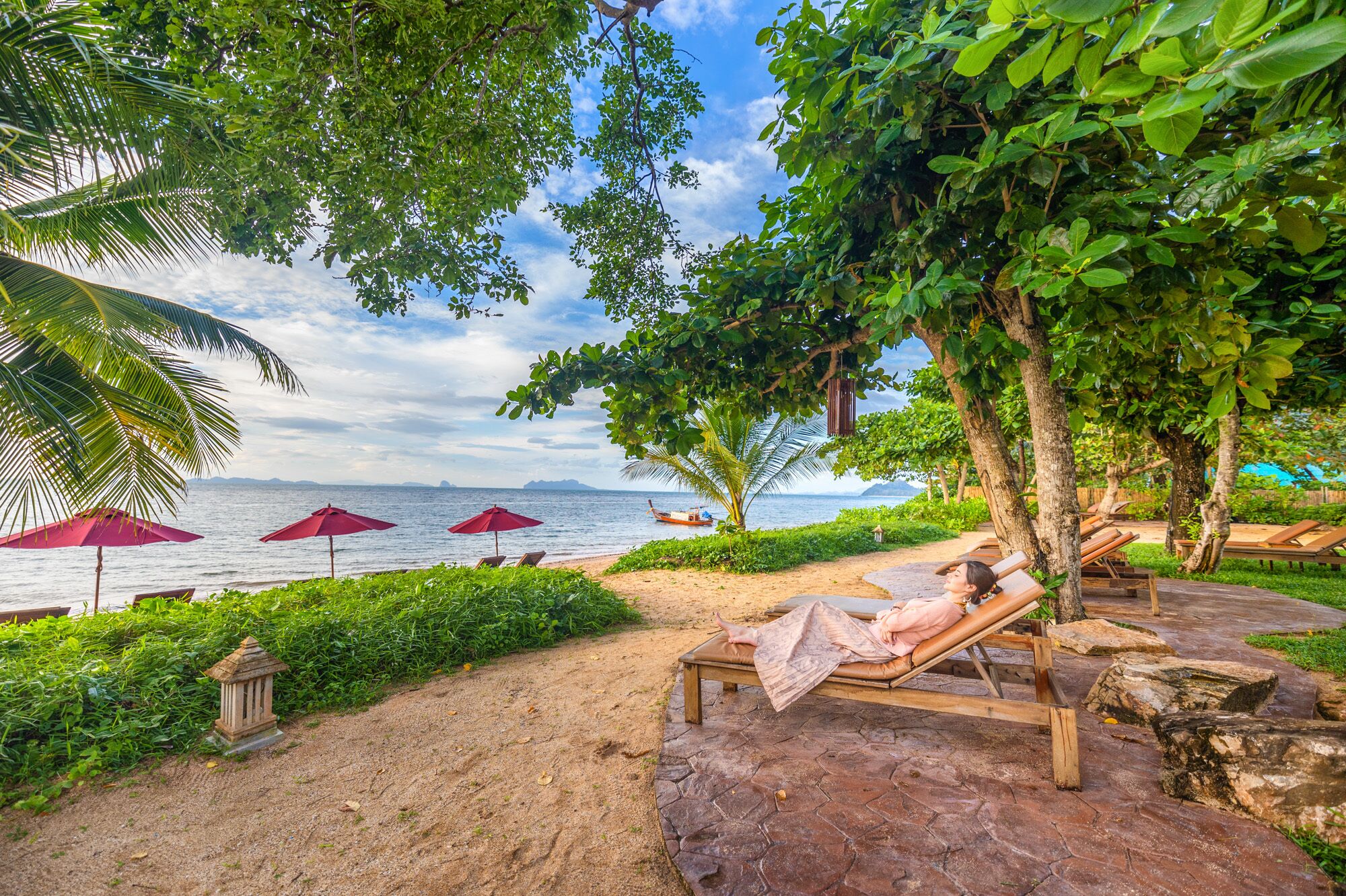 A beachside lounge area with wooden loungers, red umbrellas, and a view of the sea.