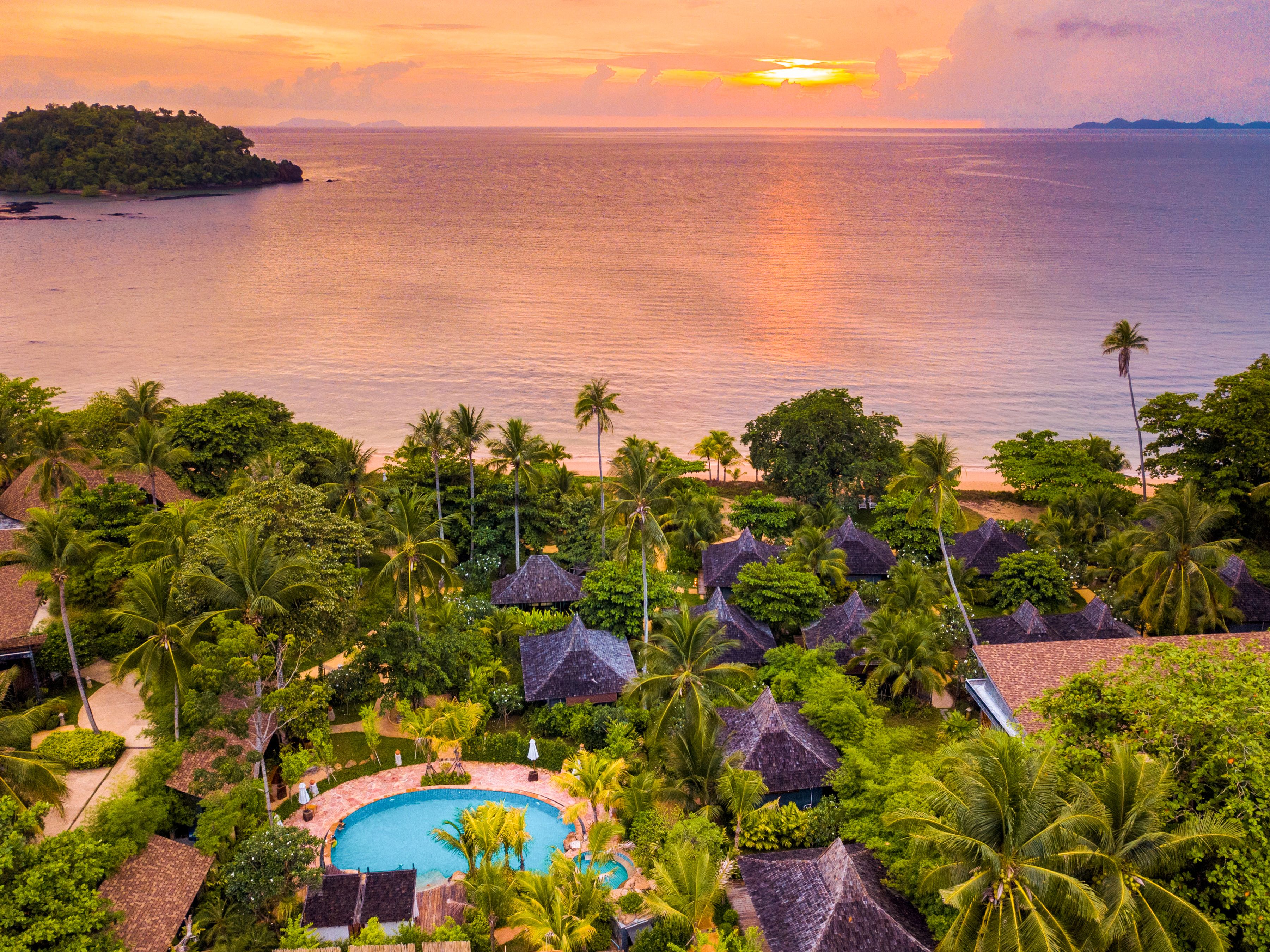 A resort with a swimming pool surrounded by palm trees and thatched-roof huts, overlooking a calm sea at sunset.