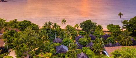 A resort with a swimming pool surrounded by palm trees and thatched-roof huts, overlooking a calm sea at sunset.