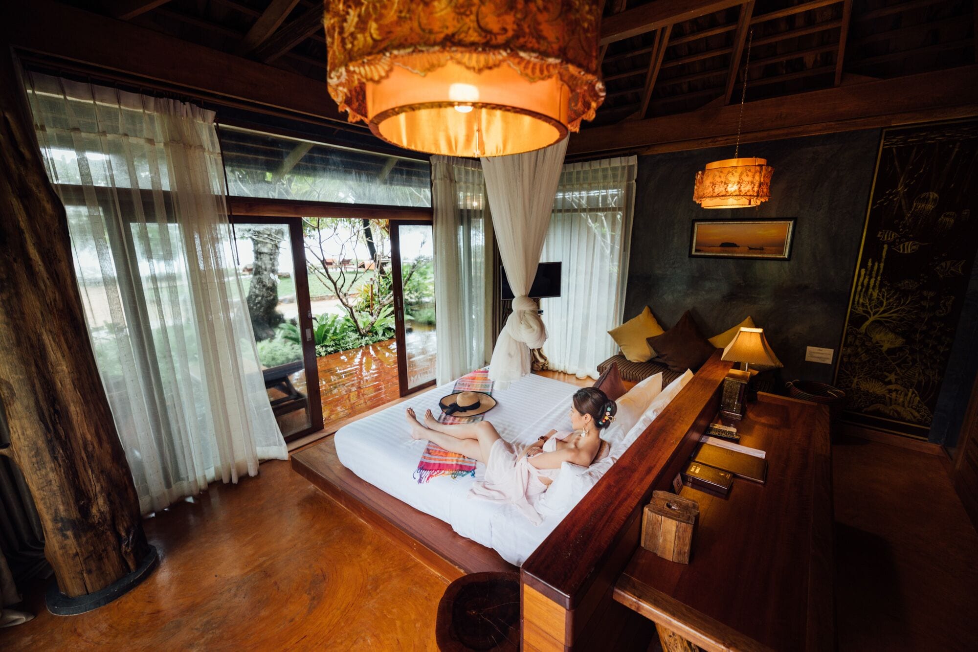 A bedroom with a large bed, a wooden floor, a hanging lamp, and a view of the outdoors.