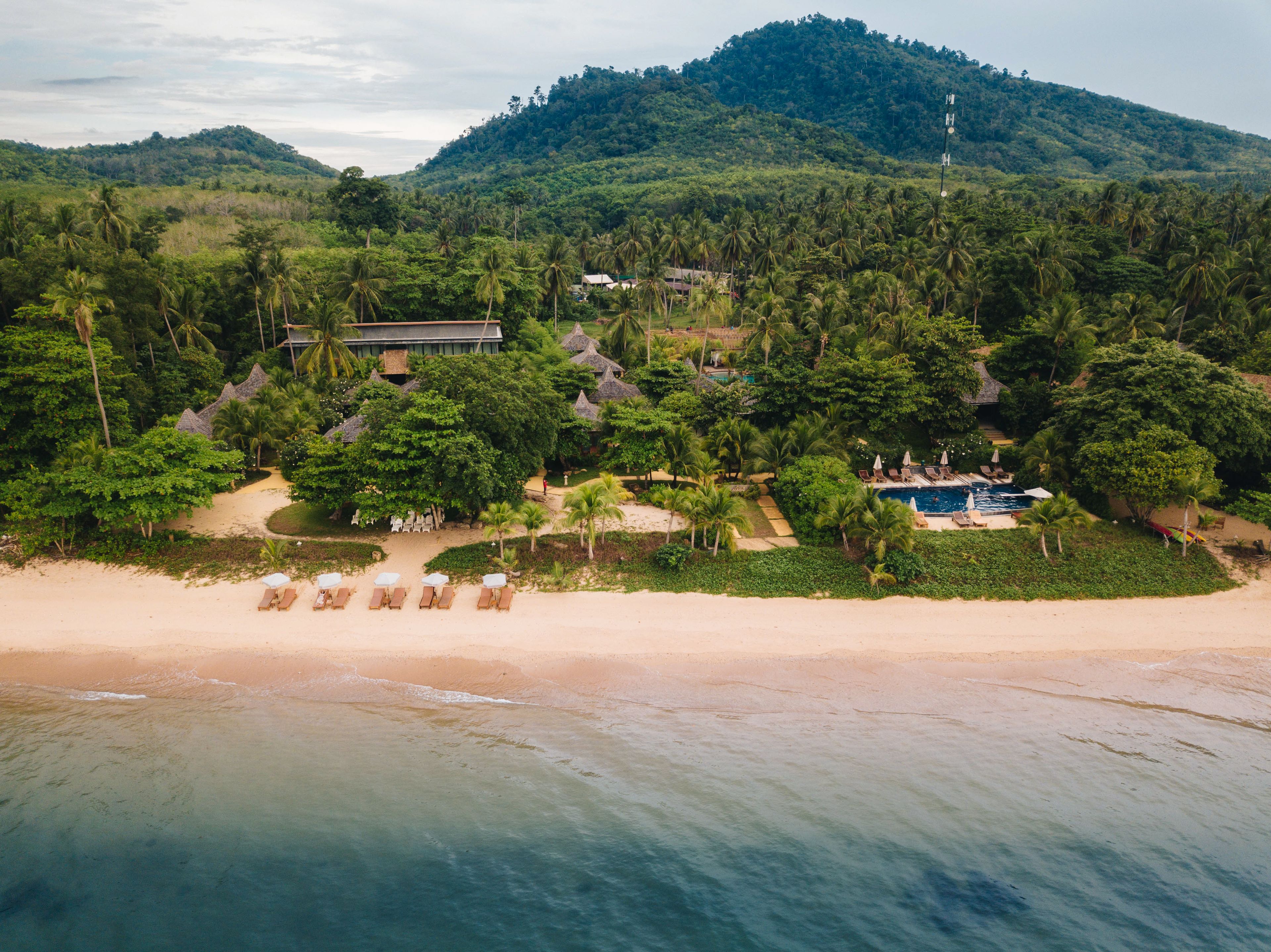 Eine Vogelperspektive zeigt einen luxuriösen Urlaubsort mit Strand, Pool und strohgedeckten Gebäuden, umgeben von dichtem tropischen Grün und Hügeln.
