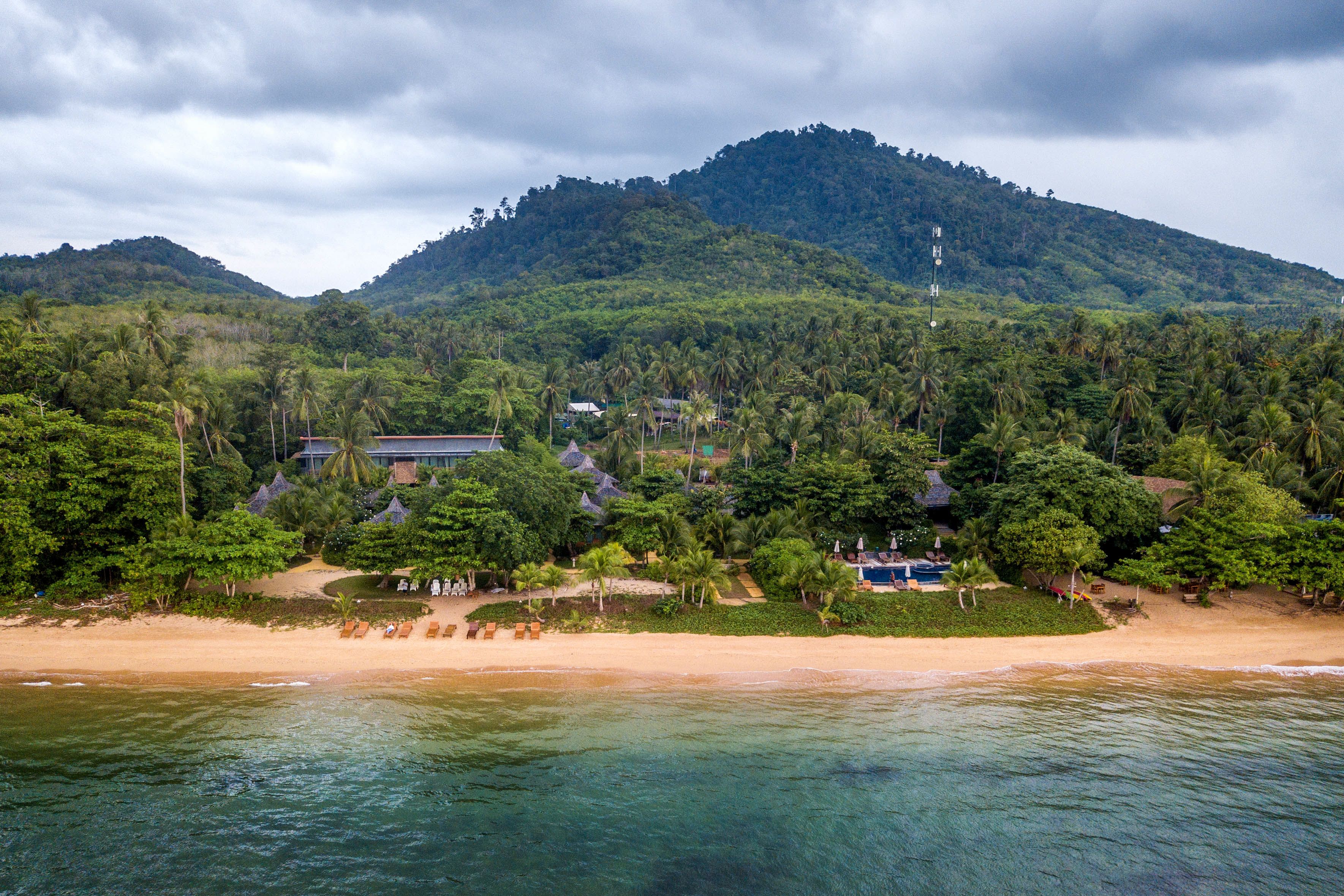 A tropical beach resort with a mountain backdrop, clear waters, and lush greenery.