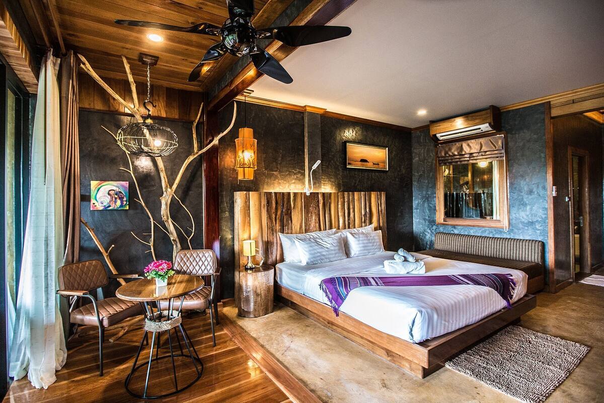 A bedroom with a bed, a chair, a table, a lamp, a ceiling fan, and a window with curtains.